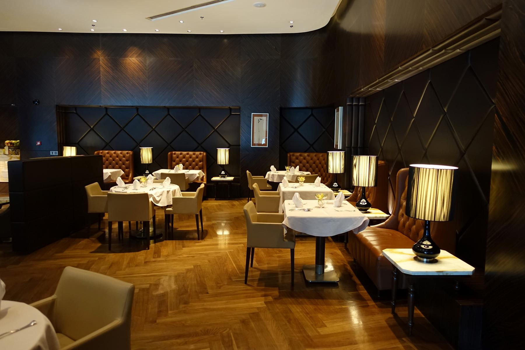 An elegant, unoccupied dining room with set tables, brown leather booths, and warm lamp lighting against dark wood walls.