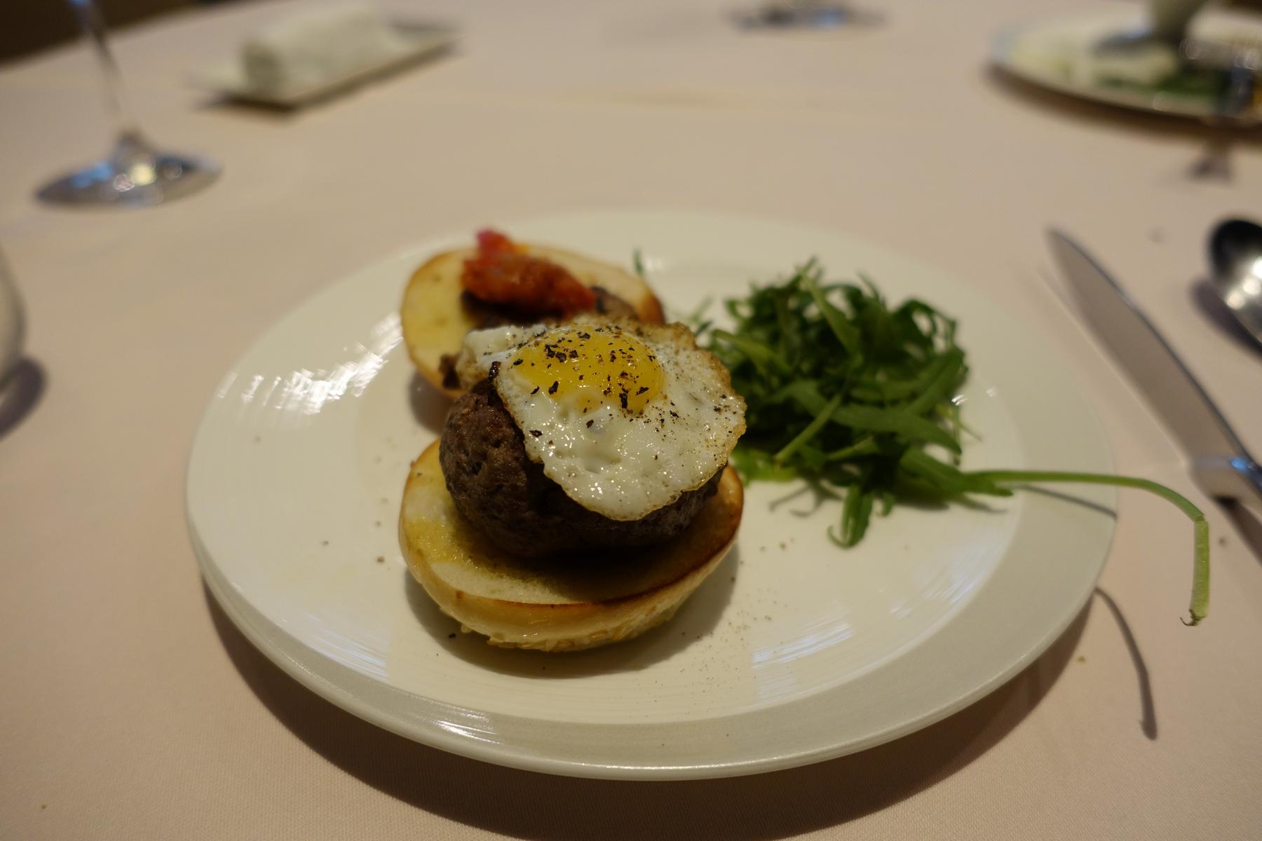 A small burger topped with a fried egg and pepper, served with arugula on a white plate.