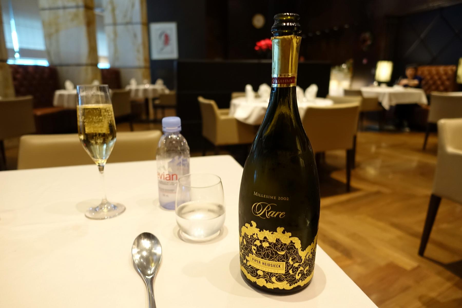 A bottle of Piper-Heidsieck Rare champagne and a glass of bubbly sit on a white table in a high-end lounge.