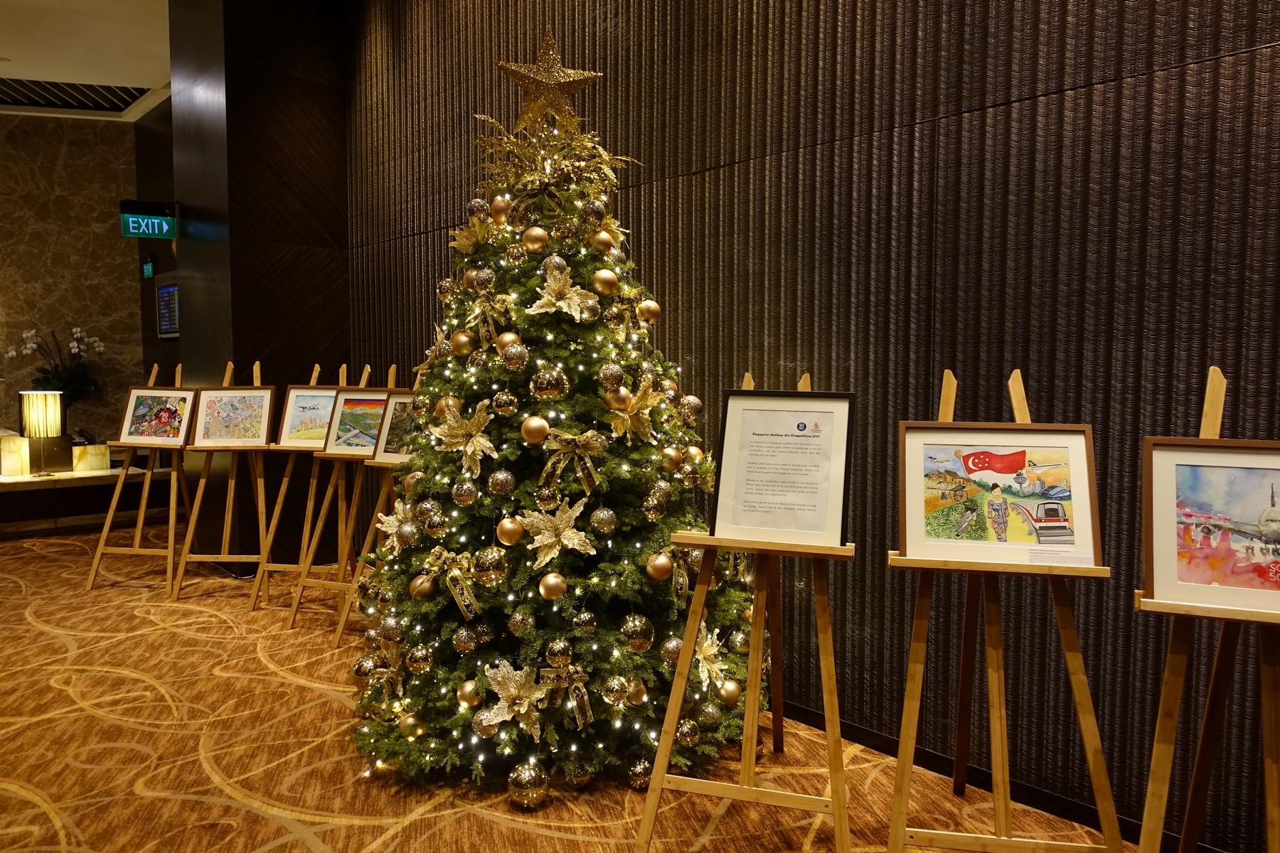 A large, gold-decorated Christmas tree stands next to an exhibit of framed art pieces on easels in a modern lounge.