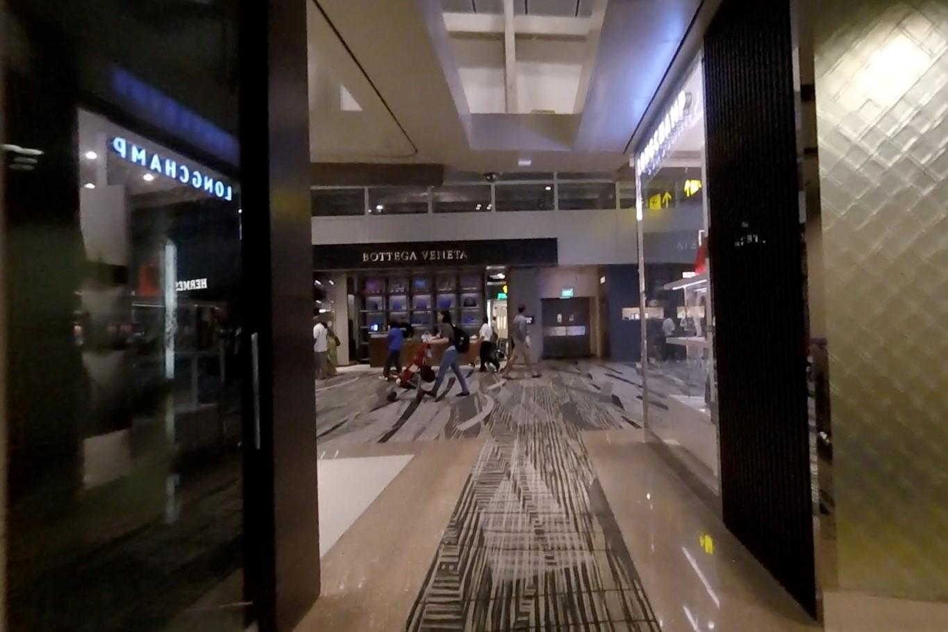 People walk past luxury stores like Bottega Veneta and Longchamp inside an airport terminal.