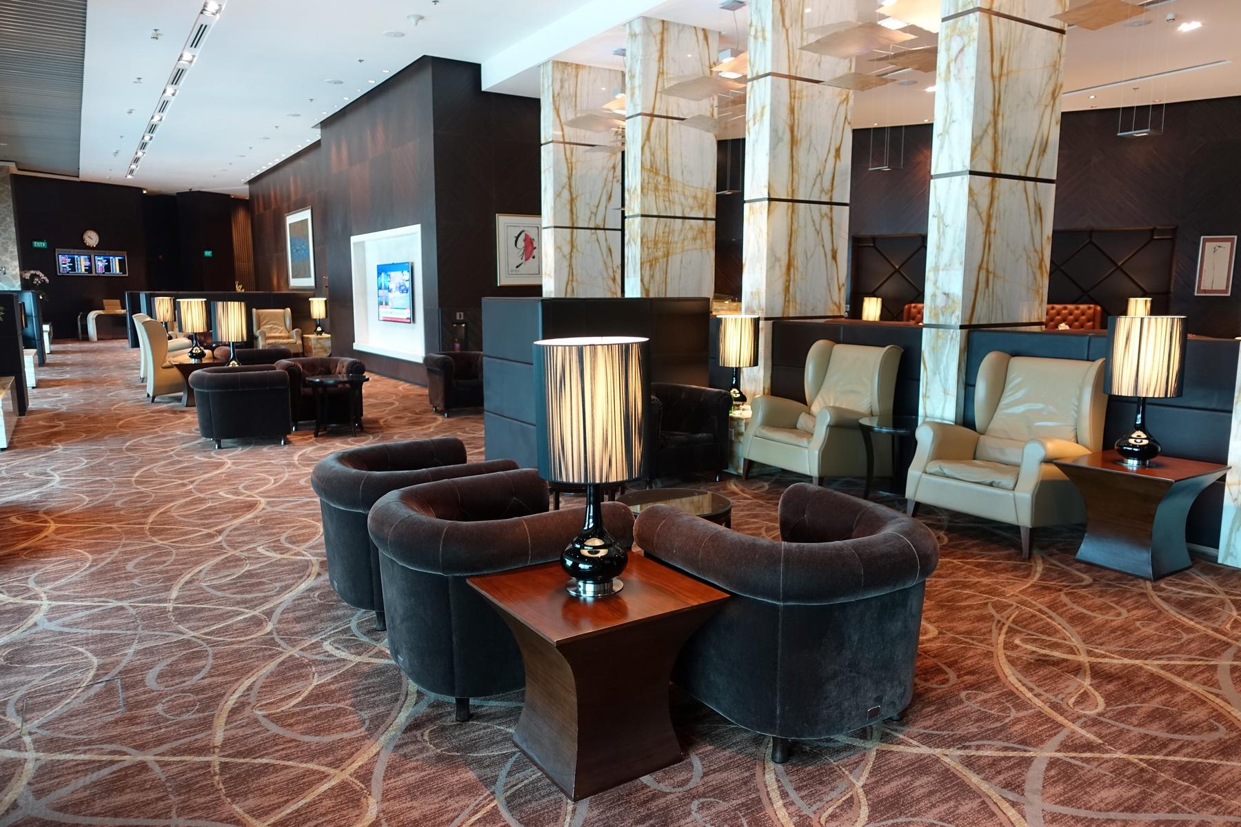 An elegant premium airport lounge with dark upholstered seating, decorative lamps, and marble-clad pillars.