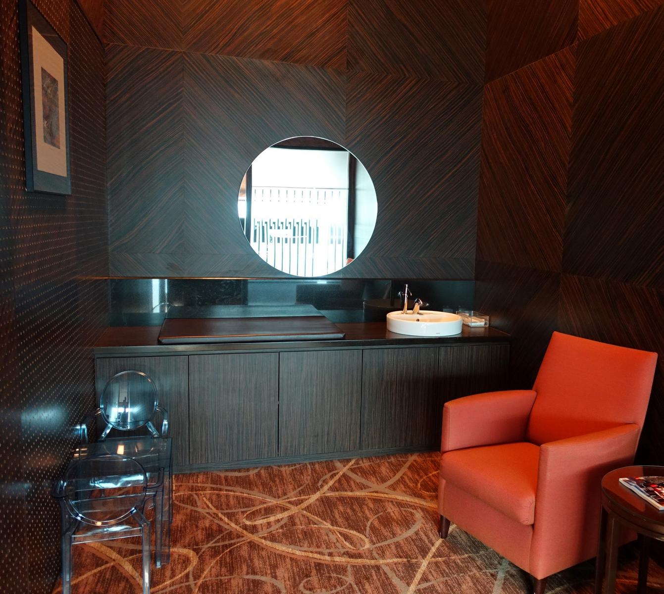 A stylish washroom area with dark wood walls, a round mirror above a sink, an orange armchair, and two clear chairs.