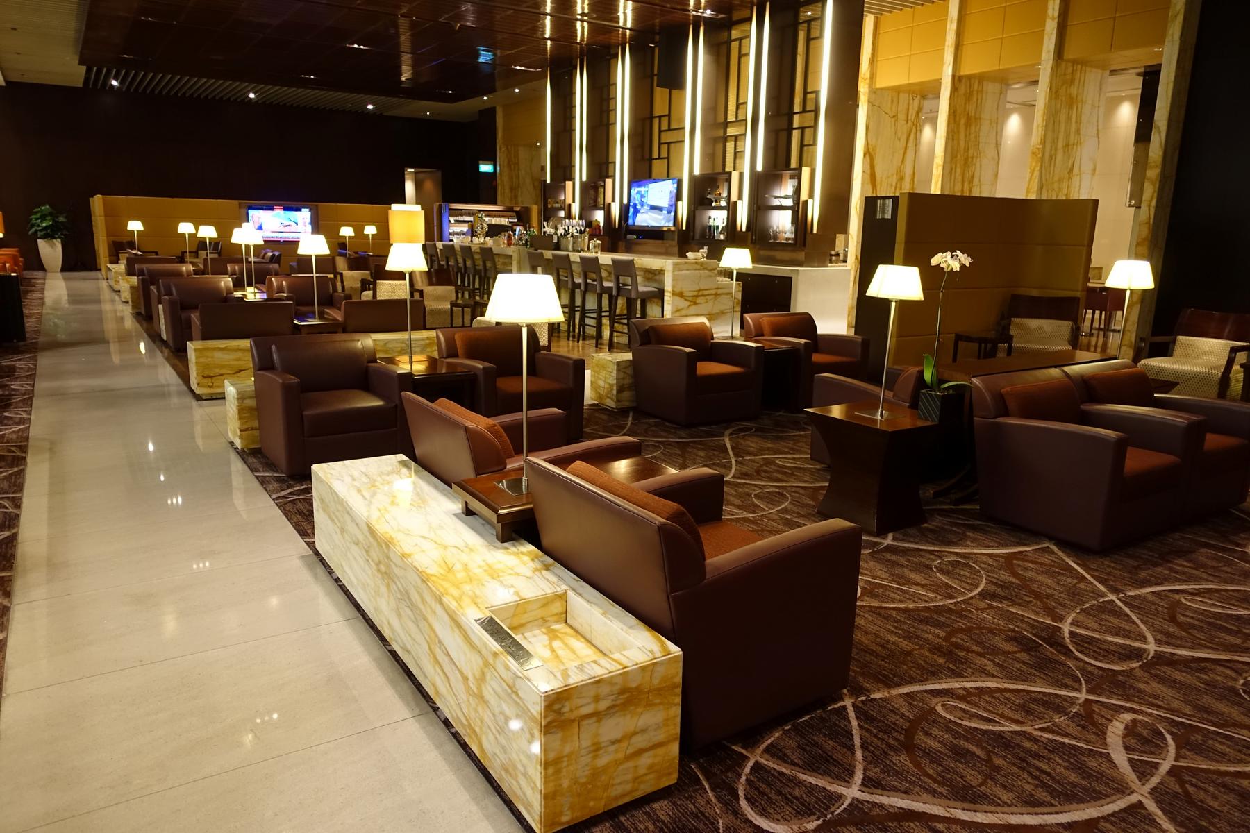 An elegant airport lounge filled with brown leather armchairs, marble tables, a long bar, and warm lighting.