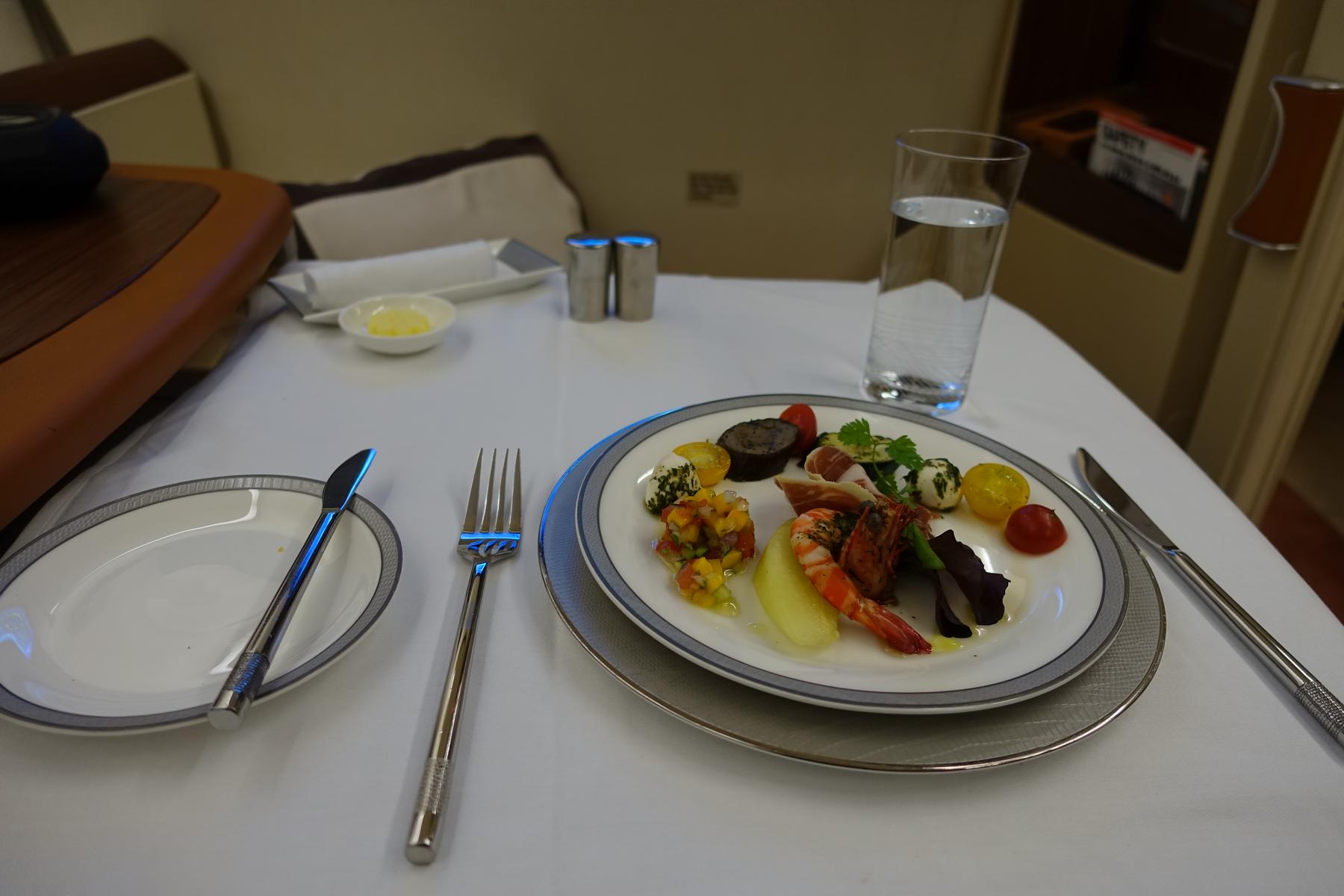 An elegantly plated appetizer featuring shrimp, cured meats, and vegetables served on a white tablecloth in an airplane cabin.