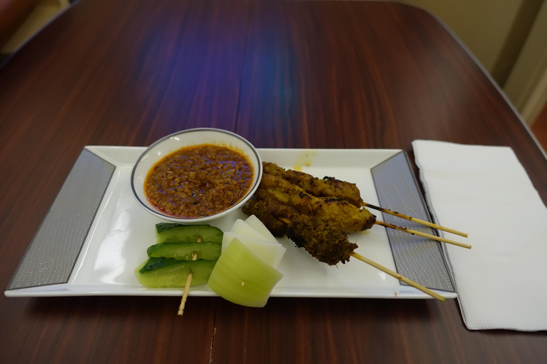 Three chicken satay skewers with peanut sauce, cucumber, and onion slices on a white plate.
