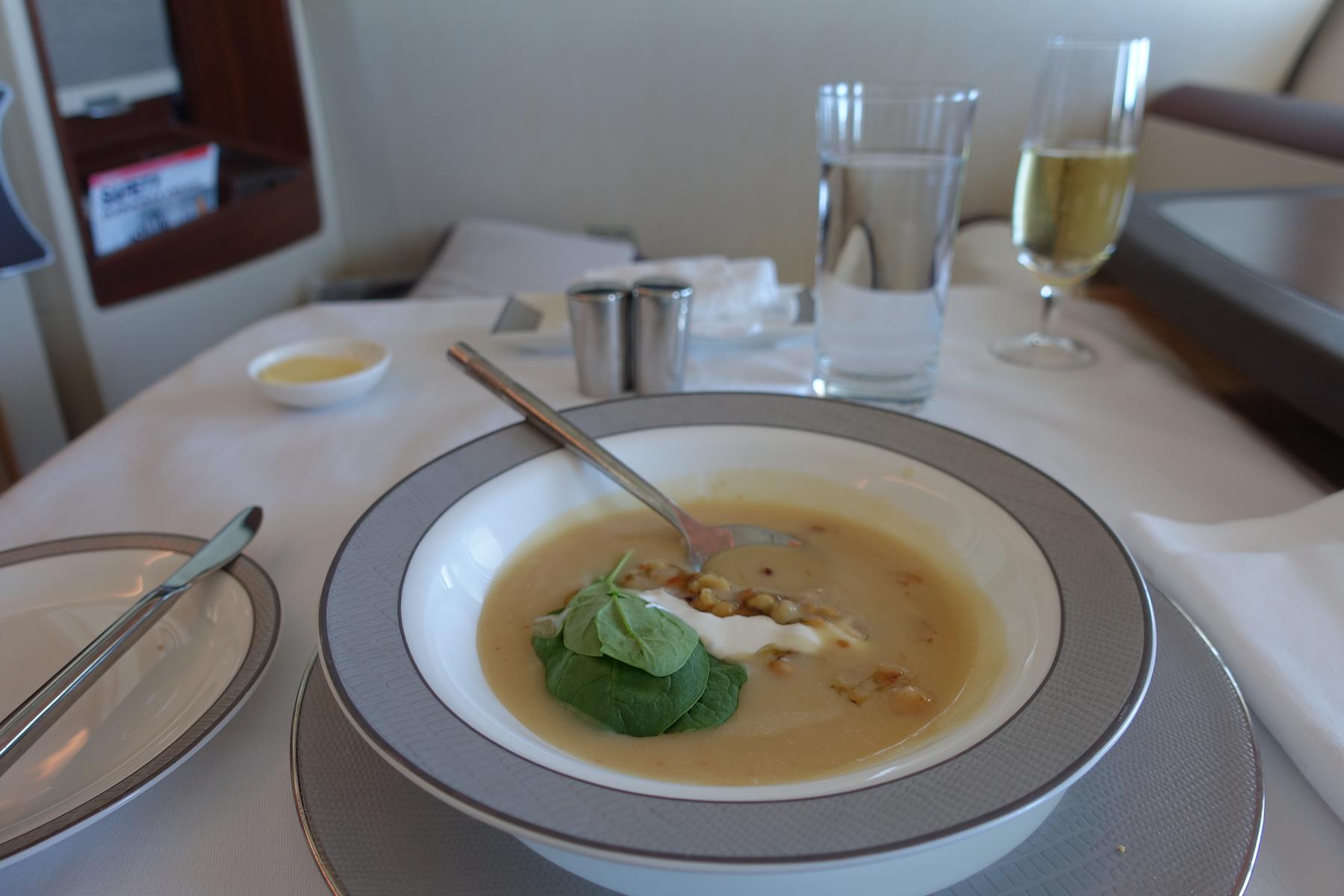 A bowl of creamy soup garnished with spinach and cream, served on a white tablecloth with drinks in an airline first class suite.