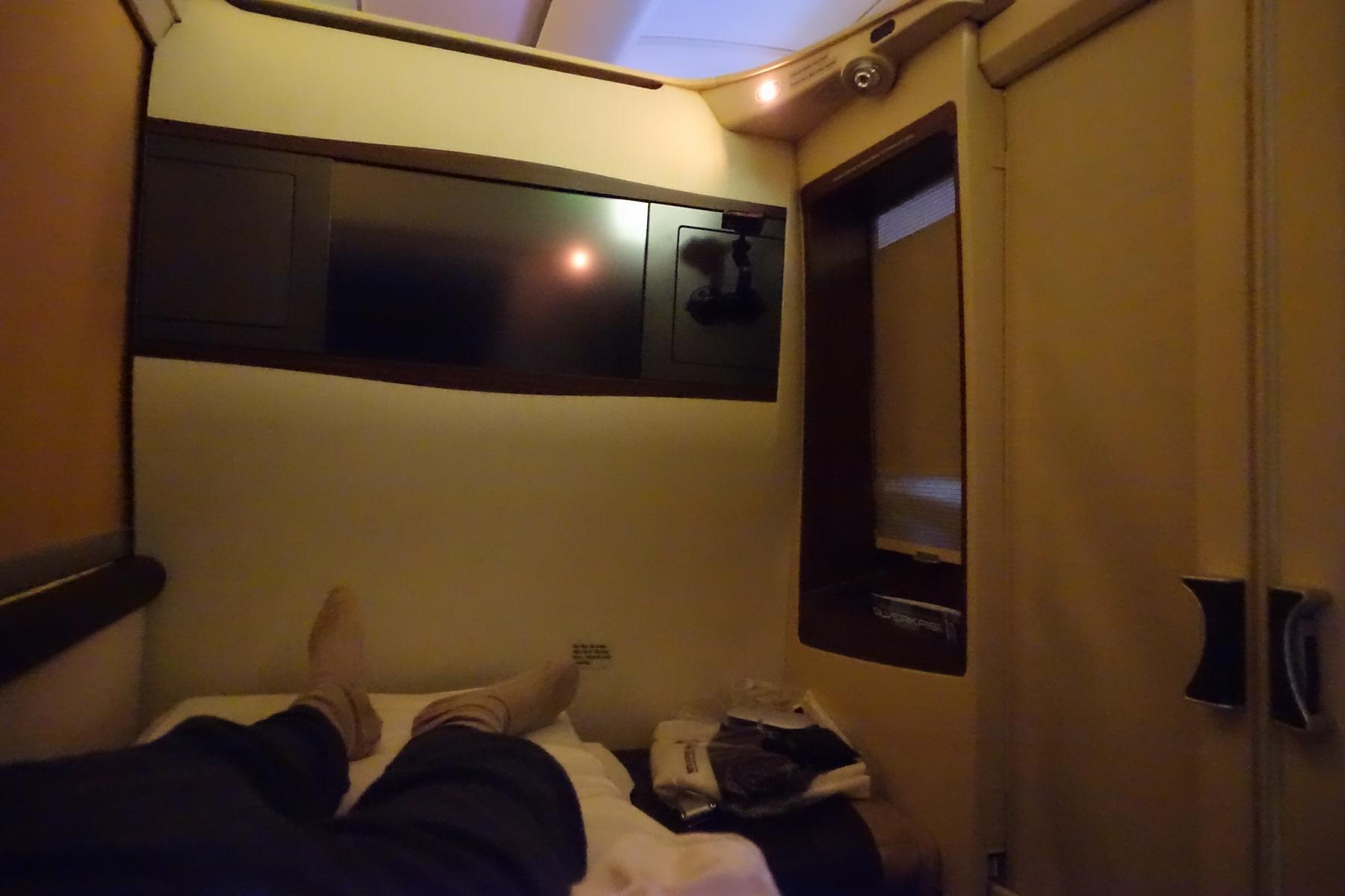View from a Singapore Airlines First Class Suite, showing a passenger's legs extended on a bed facing a large TV screen.