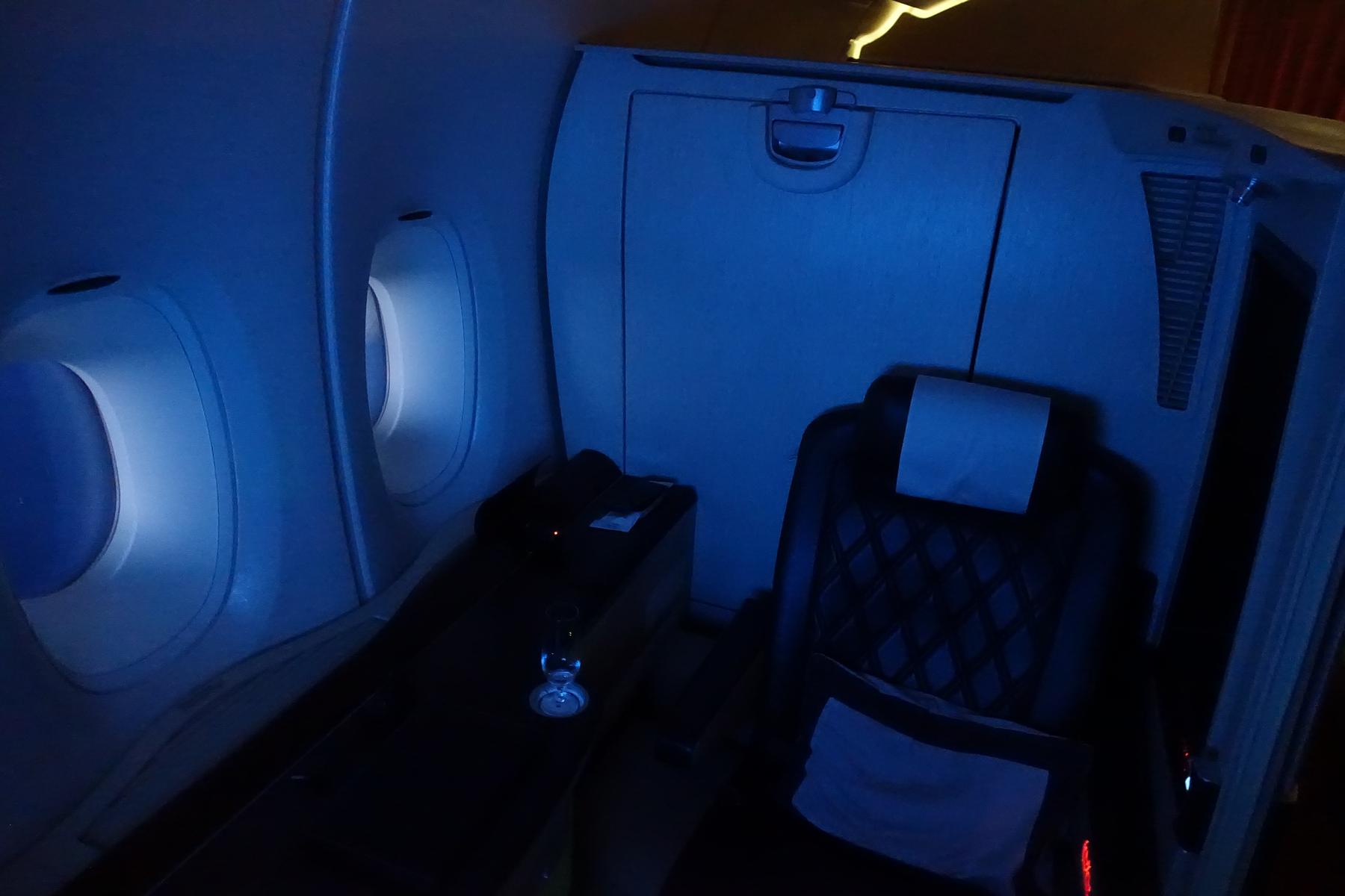 An empty Singapore Airlines First Class Suite with a dark seat, windows, and blue ambient lighting.