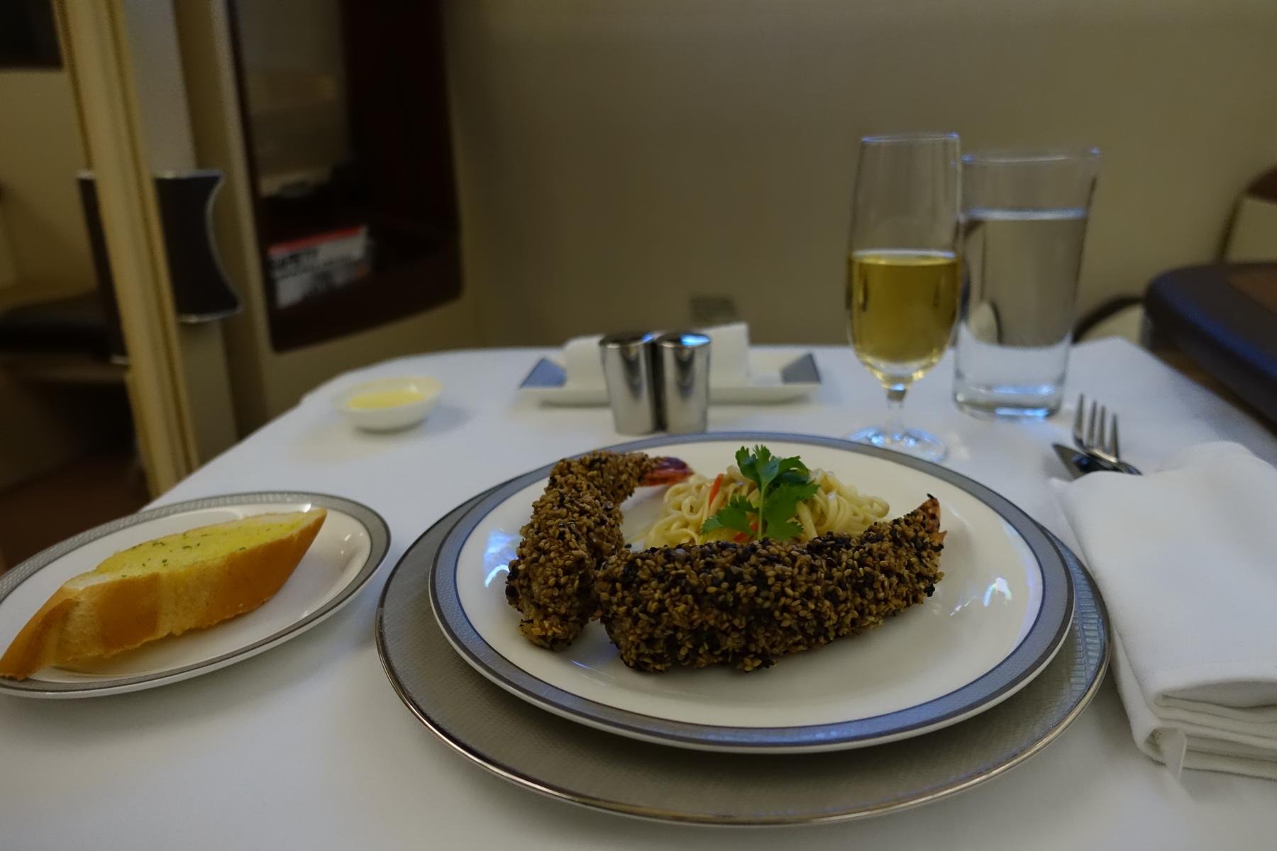 A beautifully plated airline meal features a sesame-crusted entree, noodles, garlic bread, a glass of wine, and water on a white tablecloth.