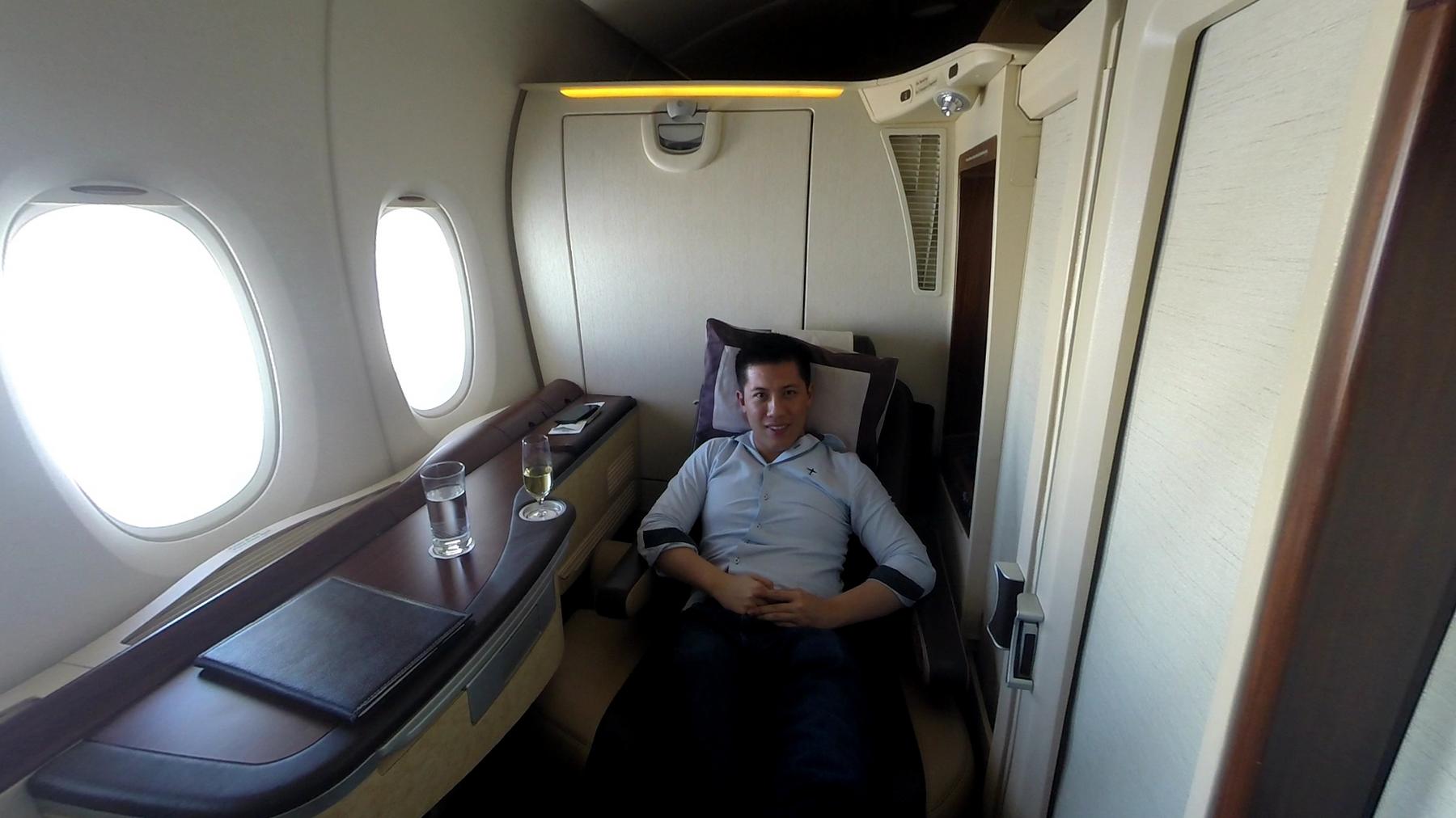 A man sits in a Singapore Airlines First Class Suite, with two windows and drinks on a side table.