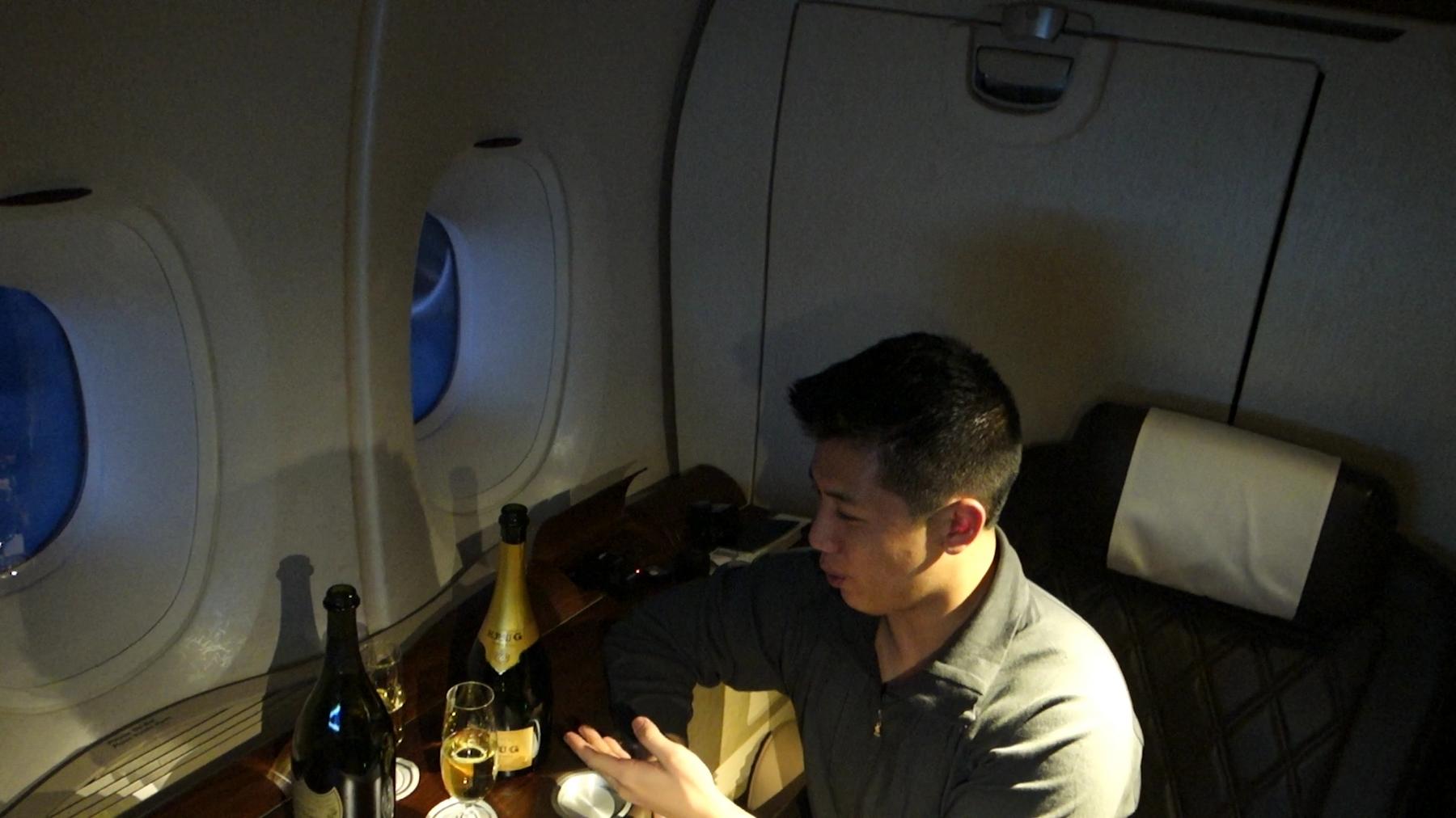 A man sits in a Singapore Airlines First Class Suite with champagne on a table by two airplane windows.