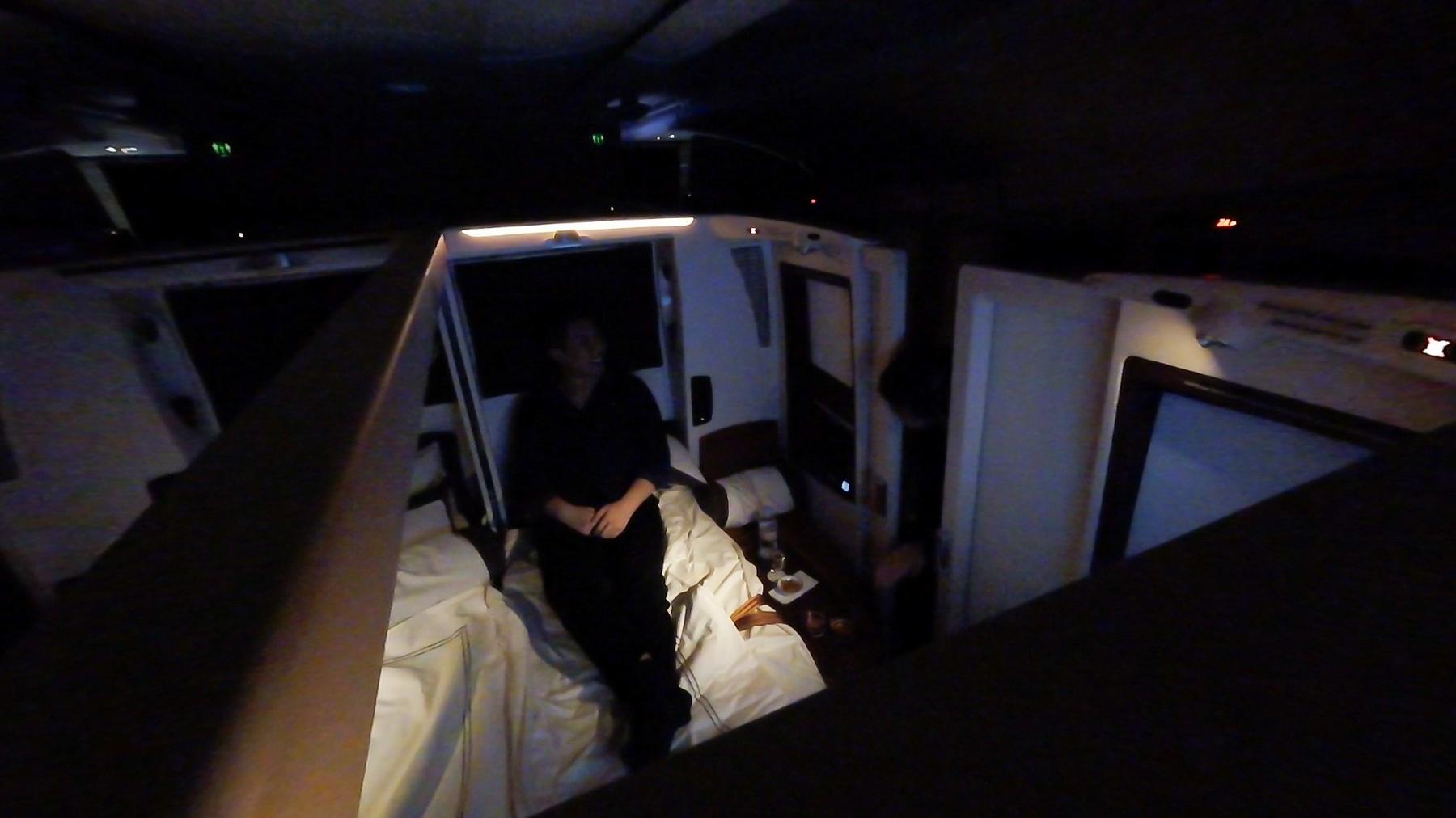 A person sits on a bed in a dimly lit, luxurious airplane suite.