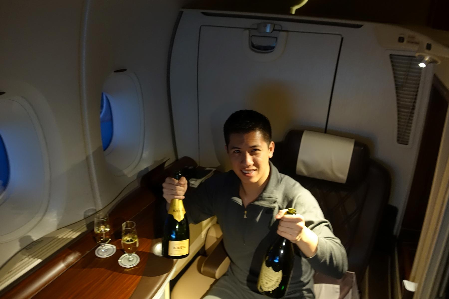 A man smiles while holding two bottles of champagne in a Singapore Airlines first class suite.