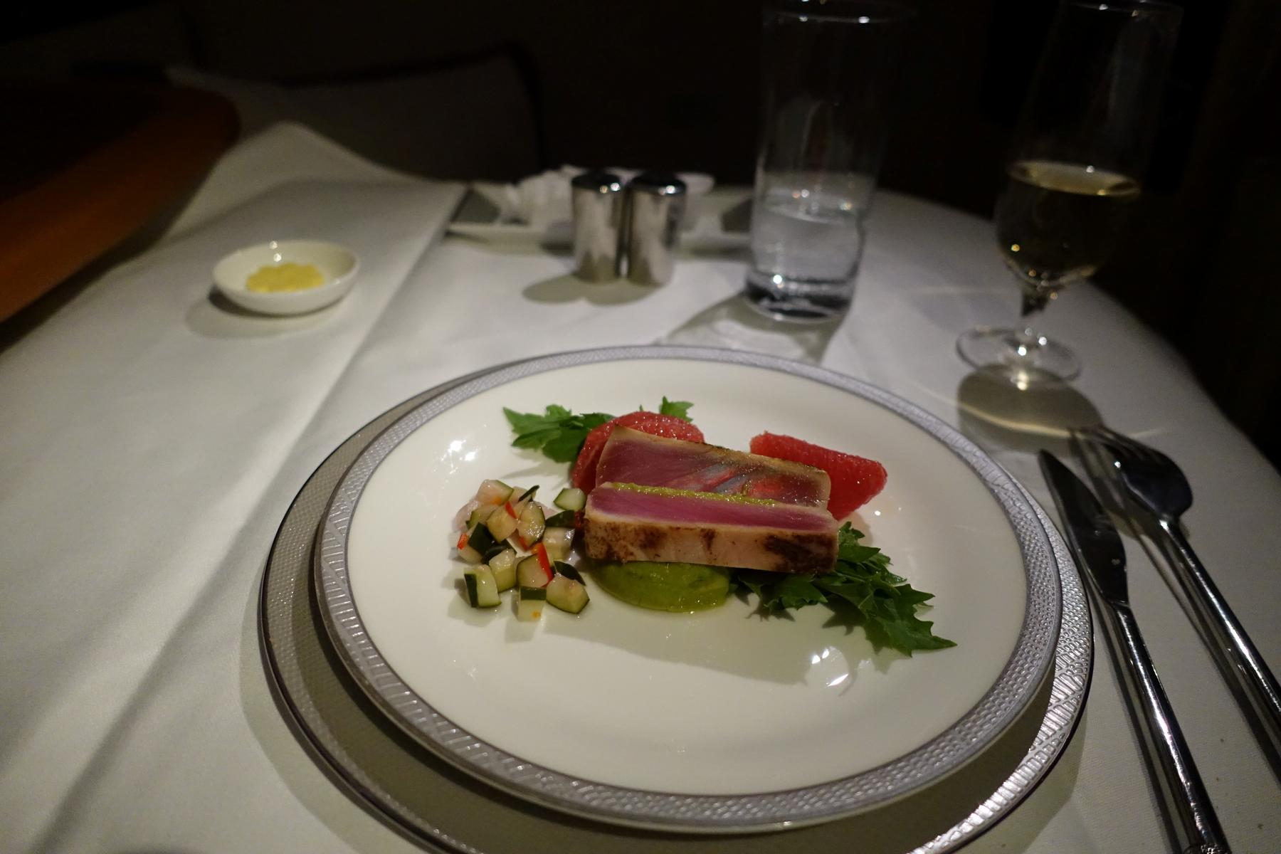A gourmet airline meal of seared tuna with grapefruit and a diced relish, served on a white plate with drinks.