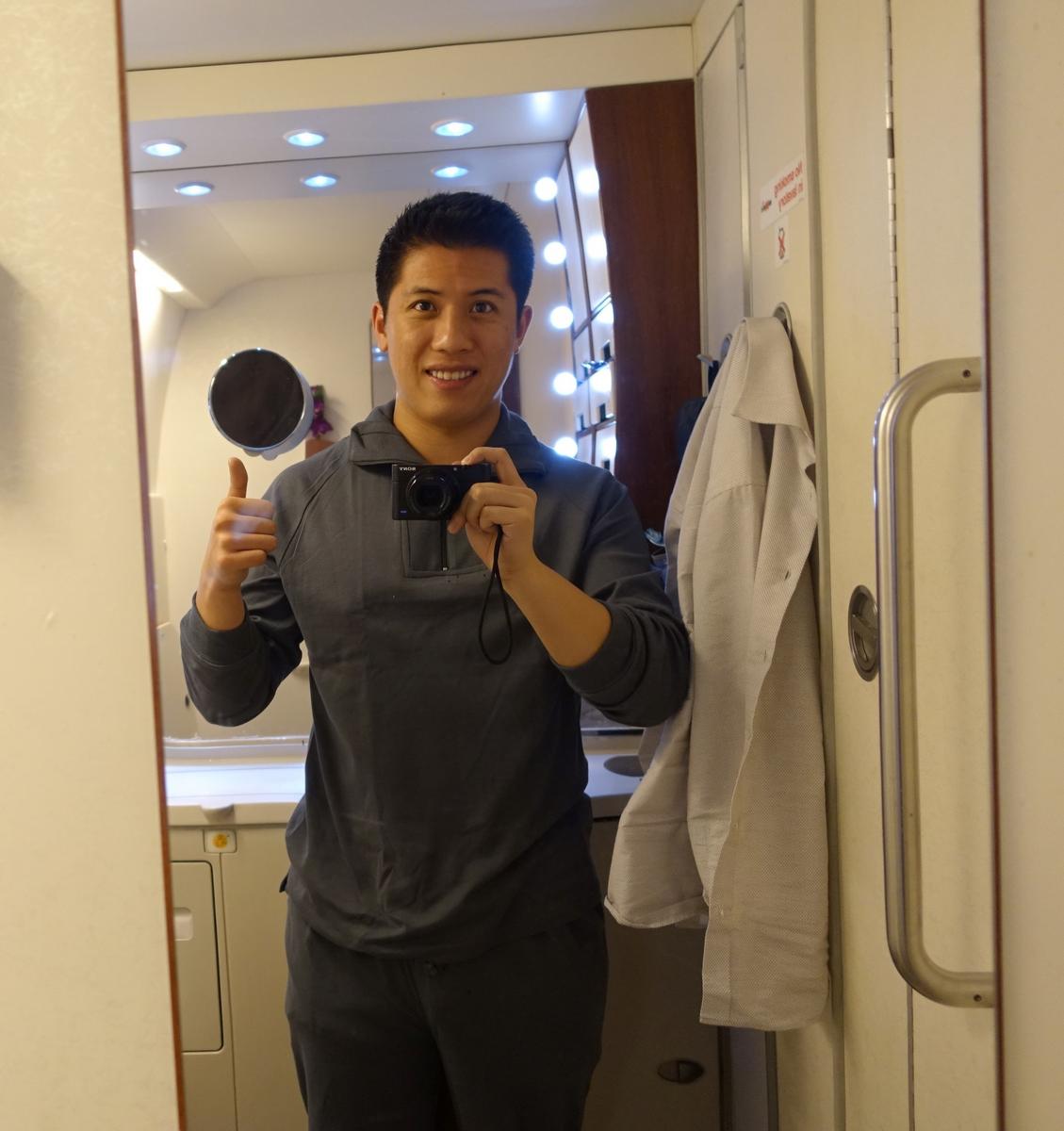A smiling man in grey pajamas gives a thumbs up while taking a mirror selfie in an airplane First Class lavatory.