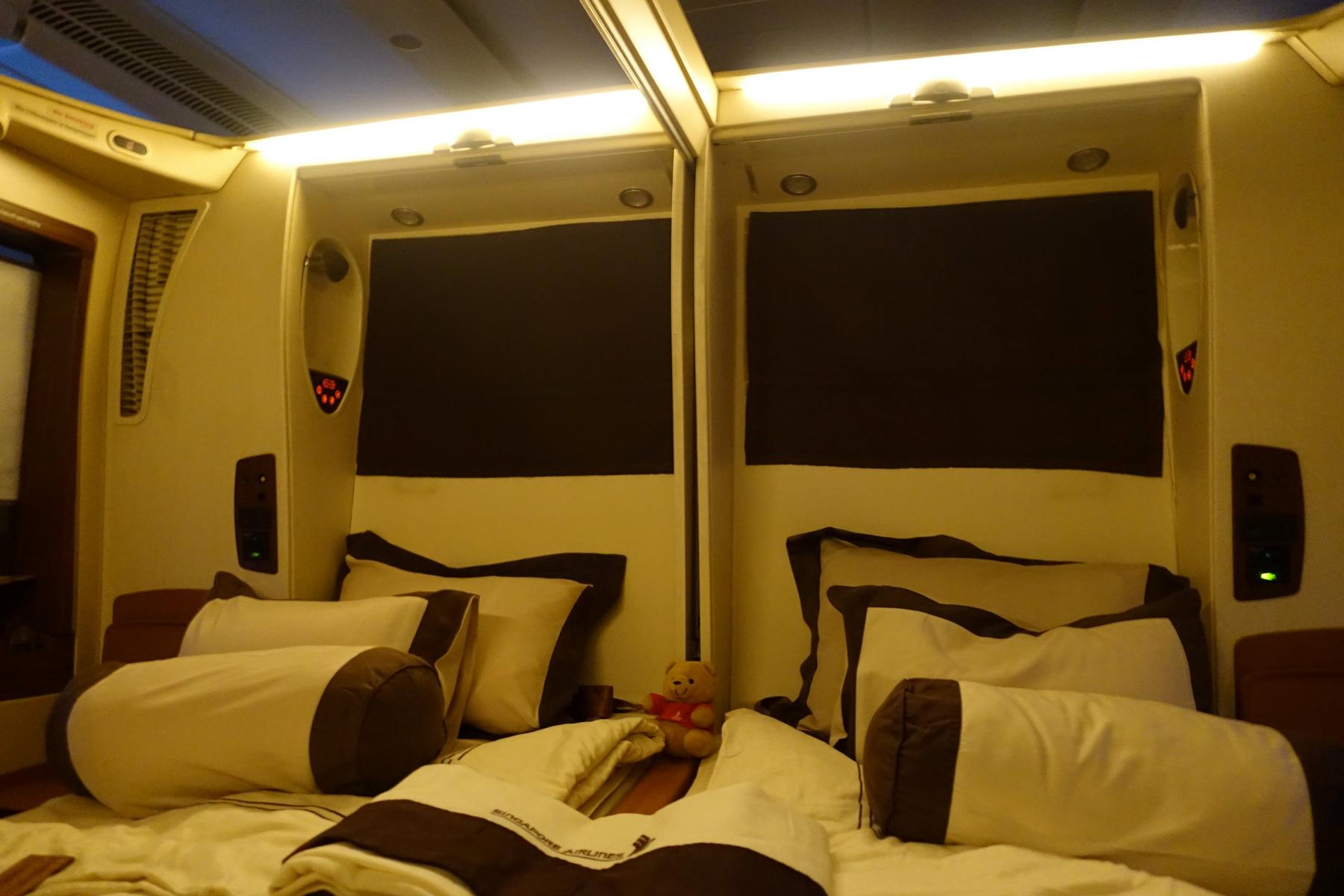 Two luxurious beds are made up in a Singapore Airlines First Class Suite, with a small teddy bear placed between them.