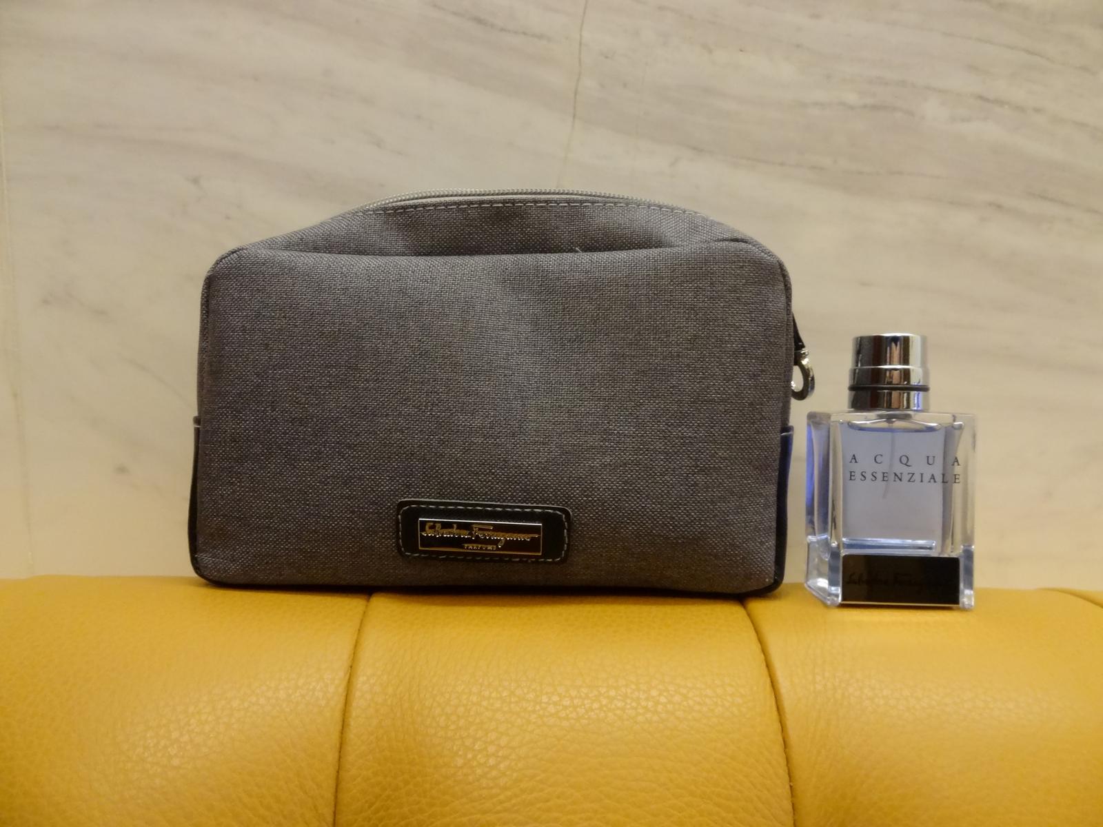 Grey Salvatore Ferragamo amenity bag and Acqua Essenziale cologne bottle on a yellow surface.