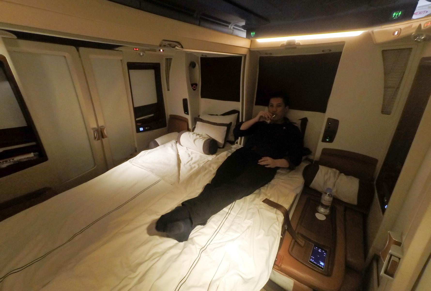 A person lies on a bed in a luxurious airplane suite, holding a drink.