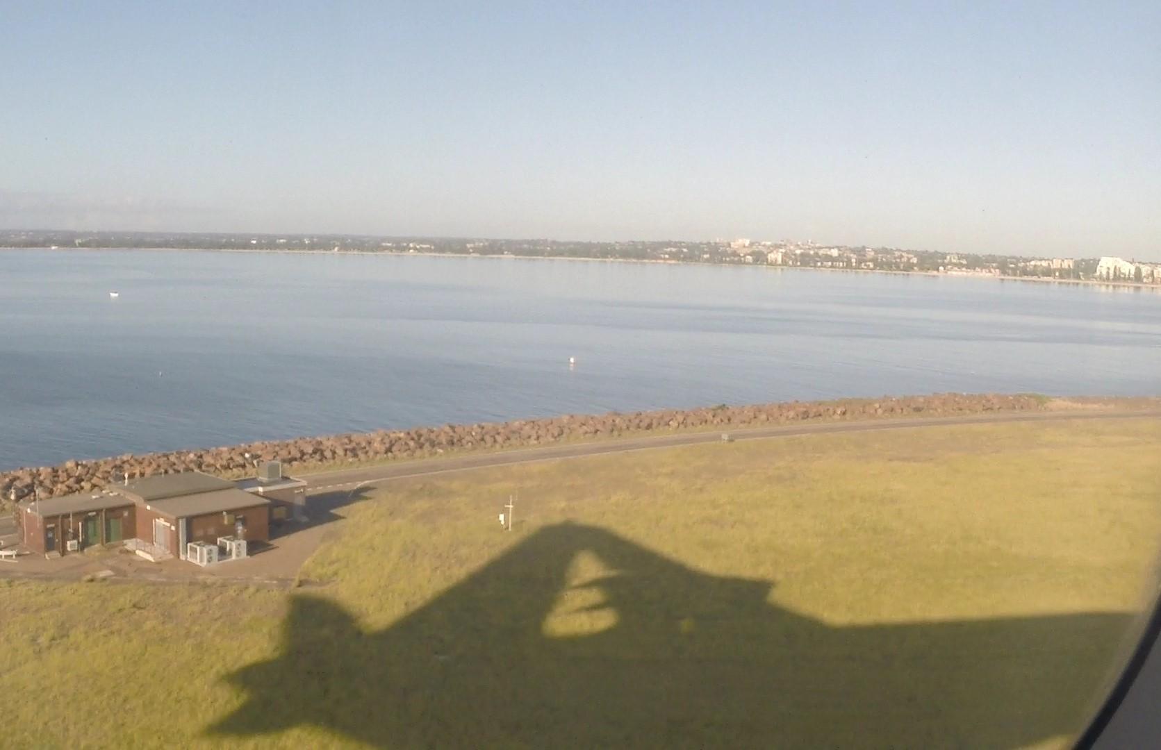 Airplane shadow on a grassy field next to a body of water with a distant city.