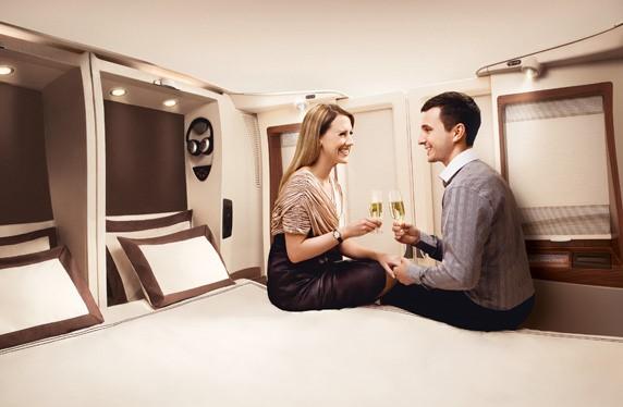 A smiling couple toasts with champagne on a bed in a Singapore Airlines First Class suite.