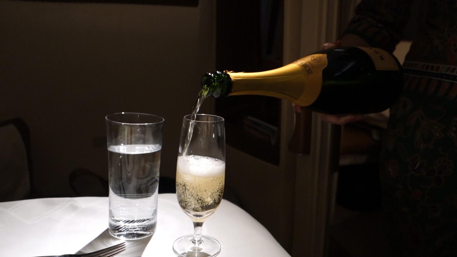 A server pours champagne from a bottle into a flute glass next to a glass of water on a table.