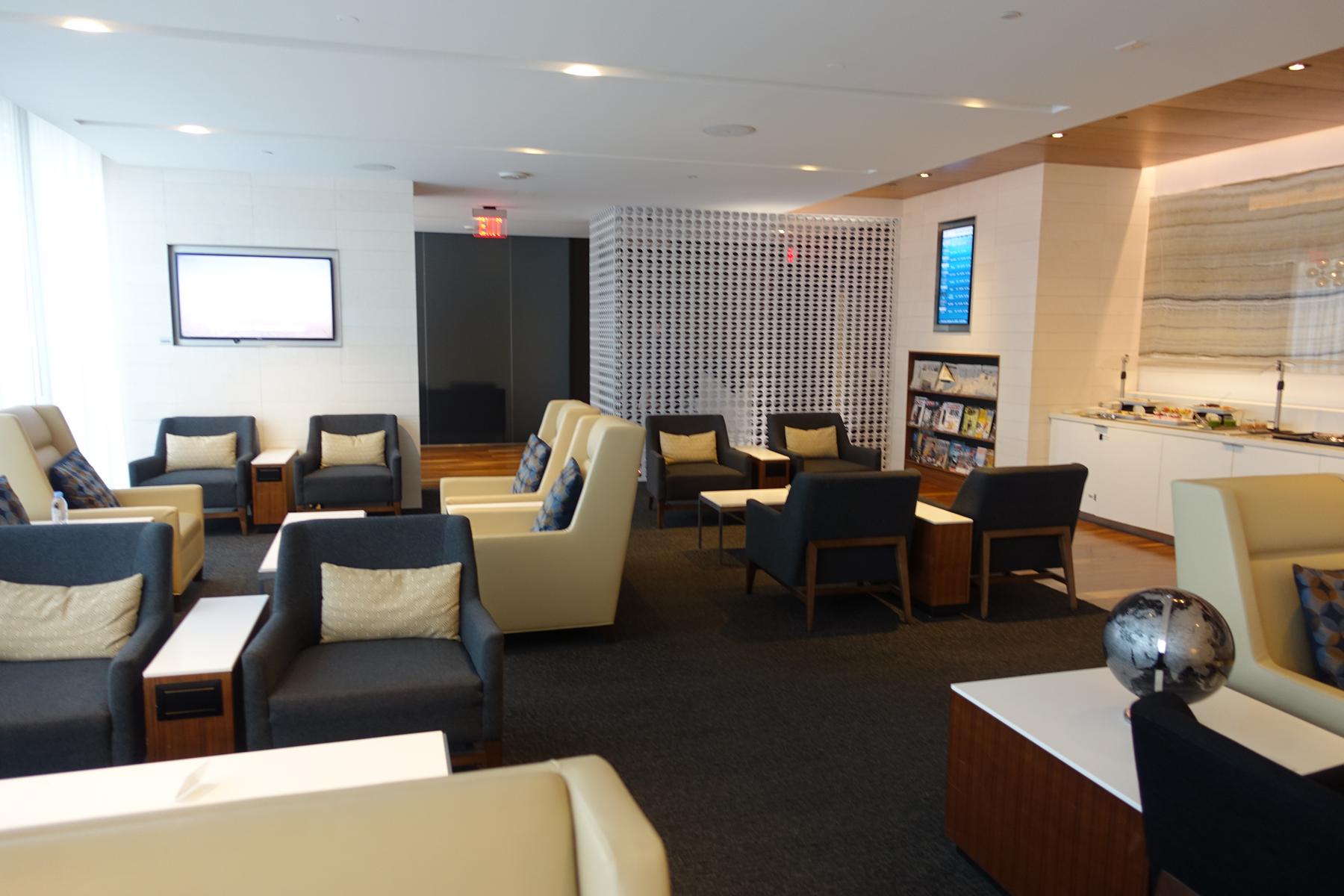 A spacious, modern airport lounge featuring various seating arrangements, a buffet station, and display screens.