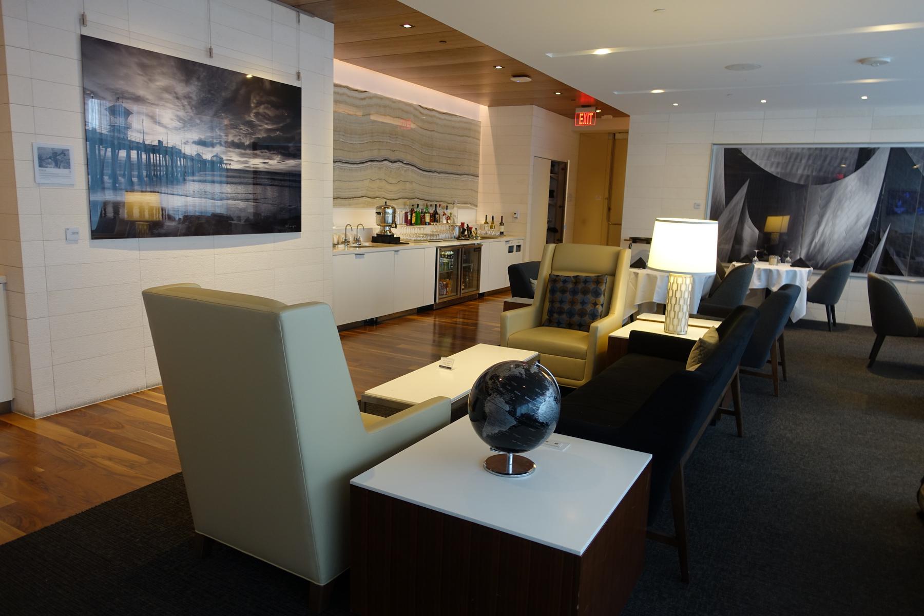 A spacious, modern airport lounge featuring comfortable seating, a well-stocked bar, and large art pieces on the walls.