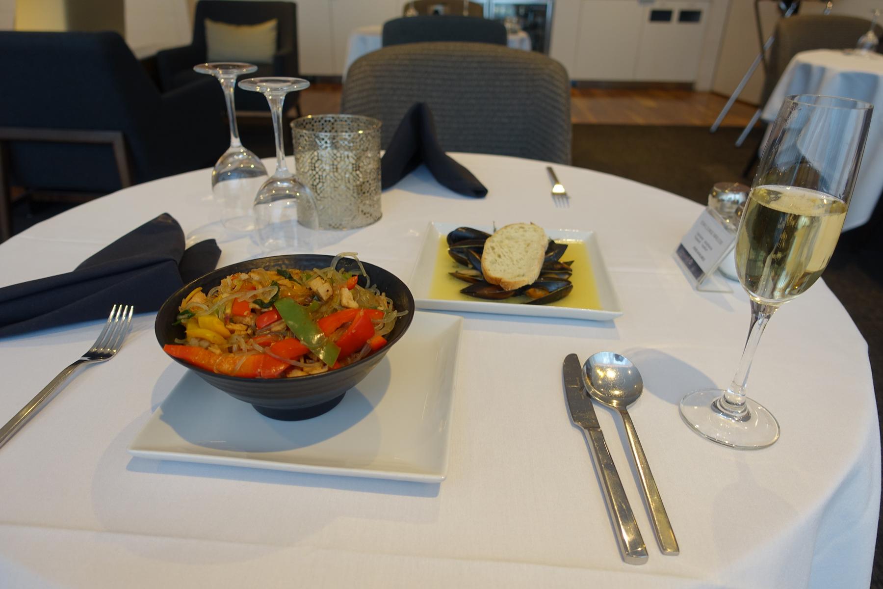 A white dining table is set with a bowl of stir-fried noodles, a plate of mussels, and a glass of champagne in a lounge.
