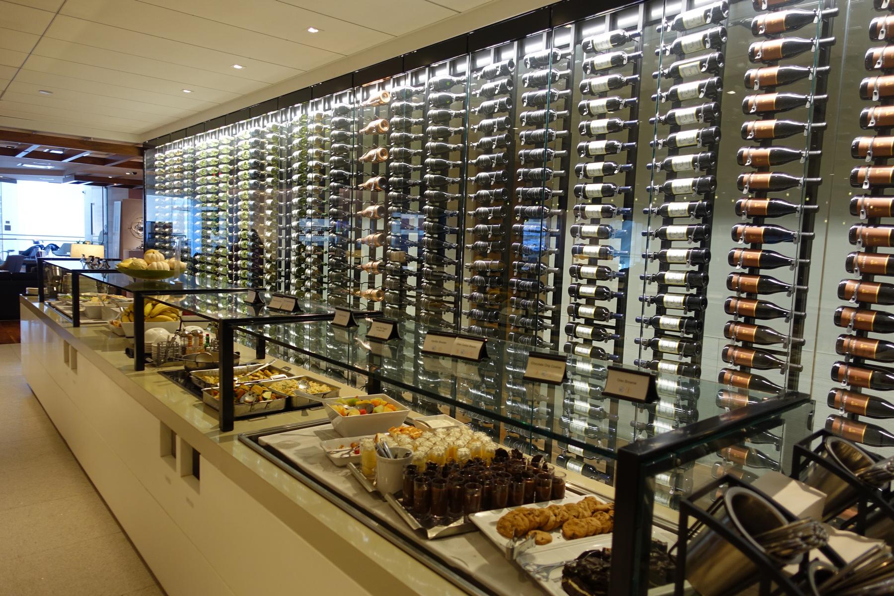 A buffet with various foods and desserts in front of a large glass wine wall.