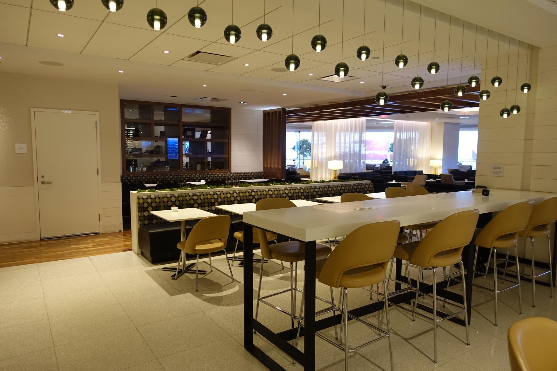 A modern airport lounge dining area with a long communal table, booth seating, and spherical pendant lights.
