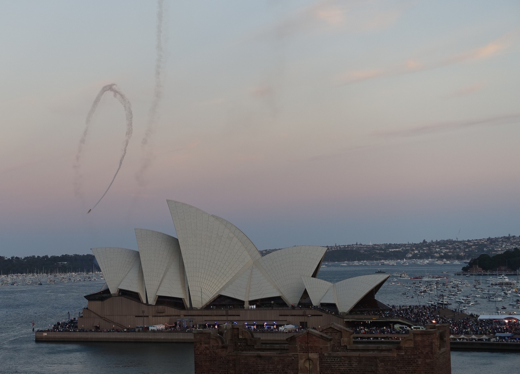 Sydney Opera House at dusk with an aerobatic plane leaving smoke trails overhead, crowds gathered, and boats in the harbor.