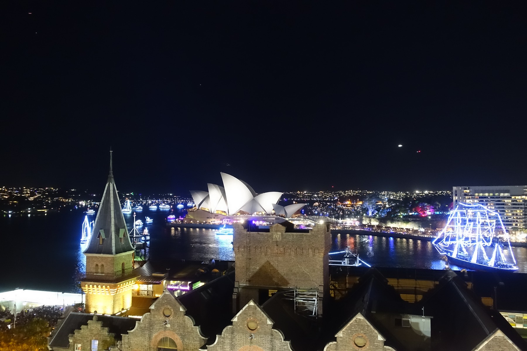 Night view of Sydney Harbour with the illuminated Opera House, festive ships, and city lights.