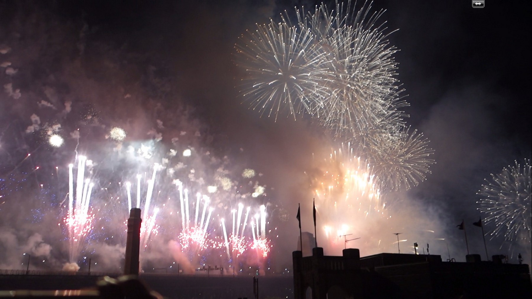 A dazzling New Year's Eve fireworks display lights up the night sky over dark building silhouettes.