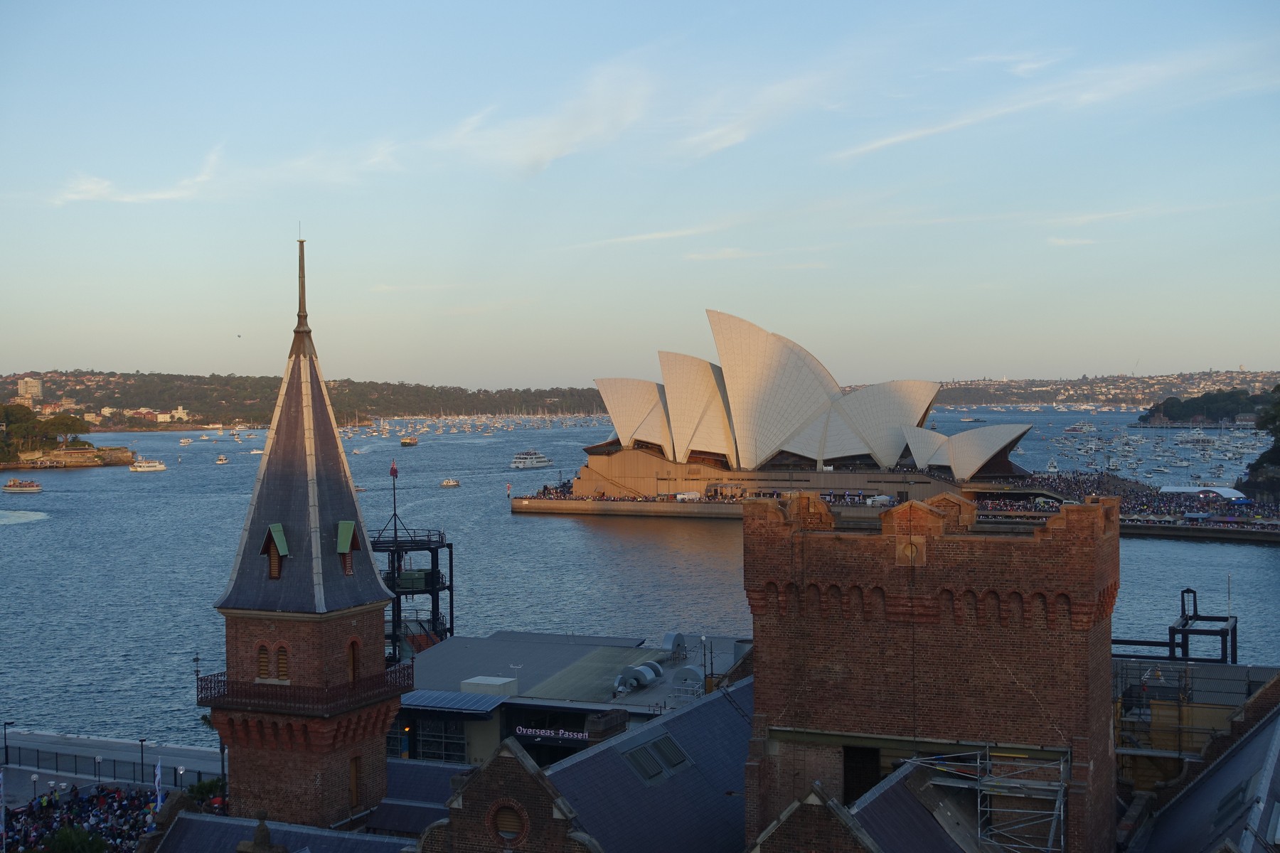 Sydney Opera House across a boat-filled harbor at sunset, with a prominent brick tower in the foreground.