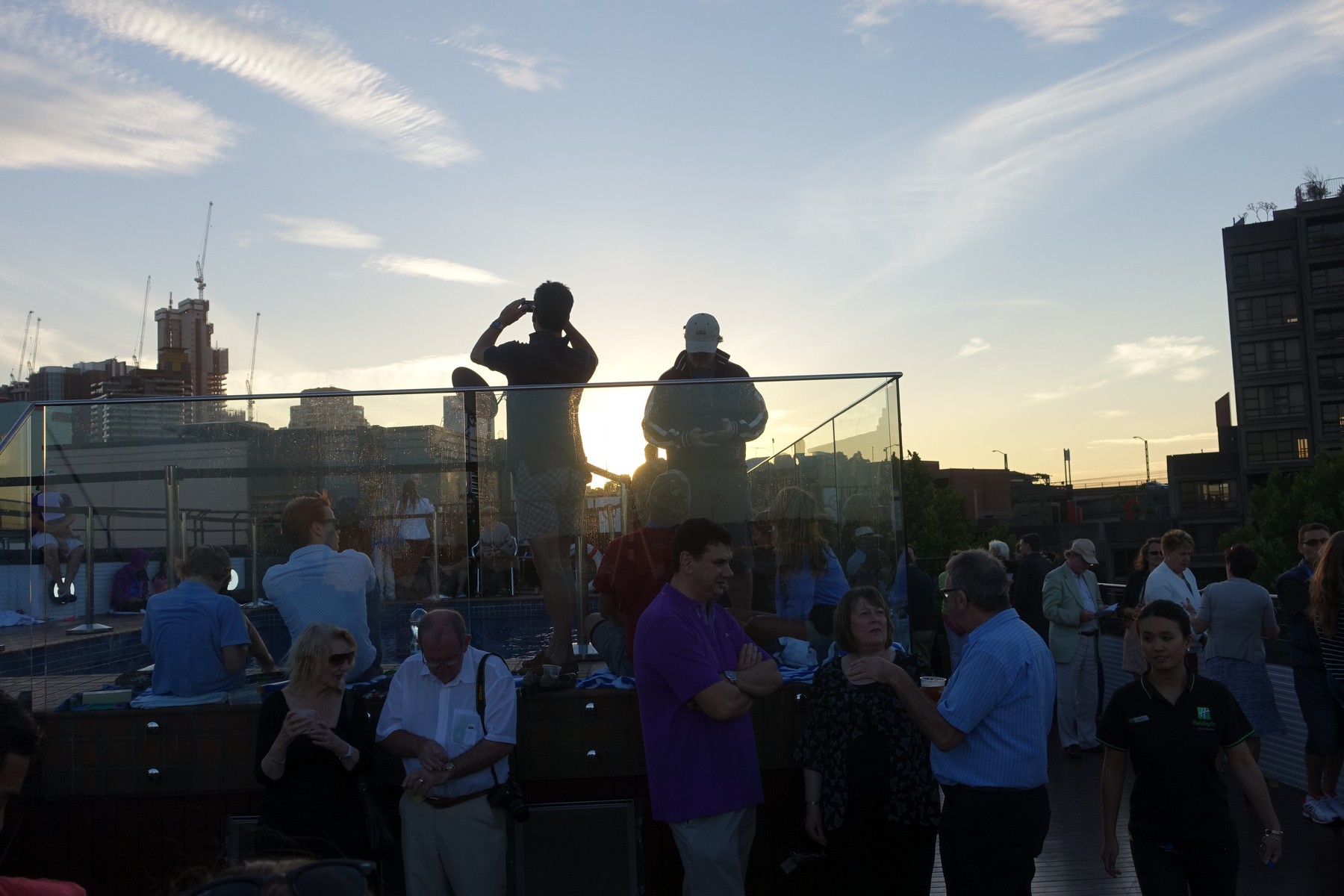 Guests gather on a Sydney Holiday Inn rooftop at sunset, with a city skyline and construction cranes in the background.