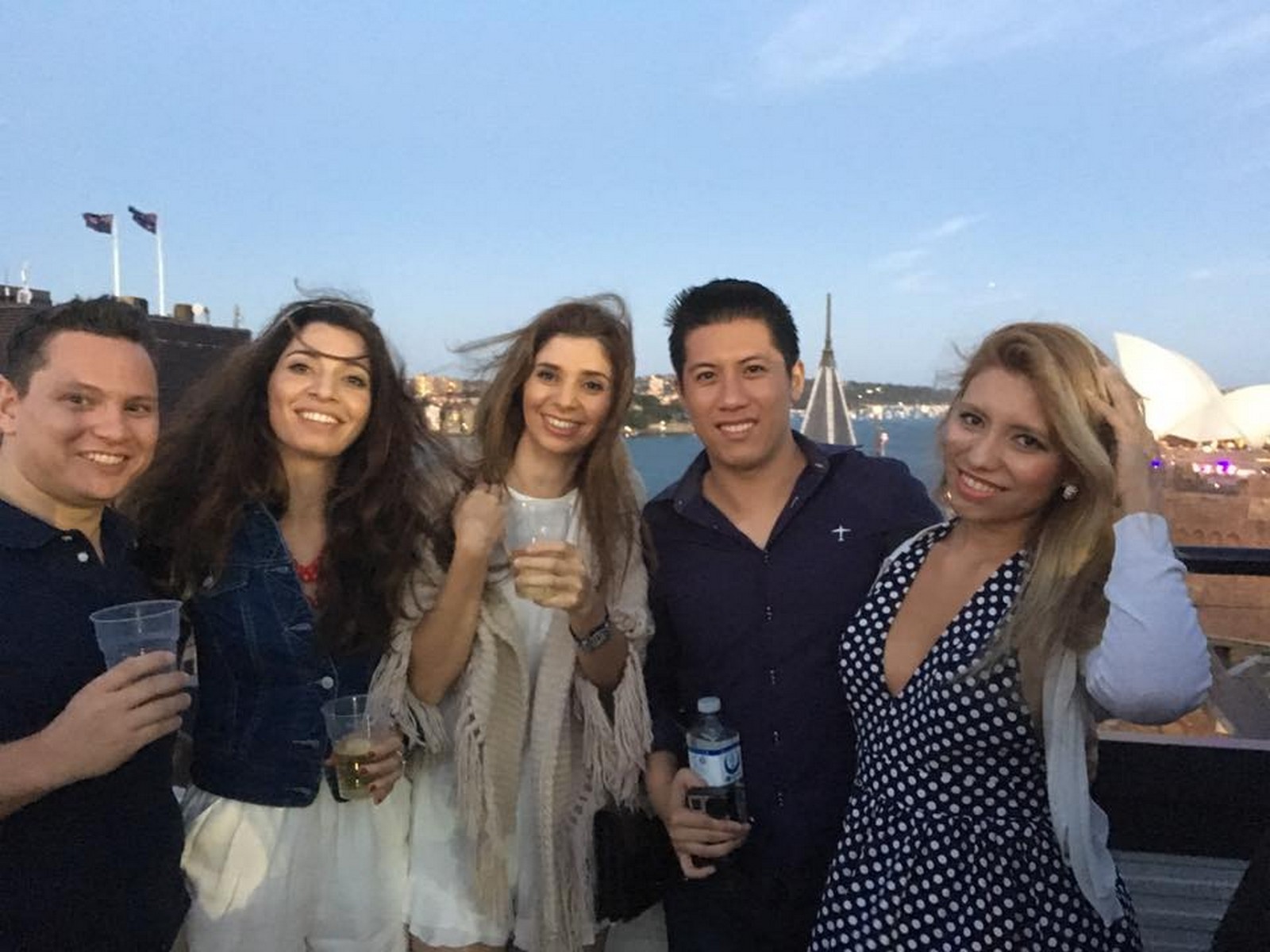 A group of five smiling people hold drinks on a rooftop overlooking the Sydney Opera House.