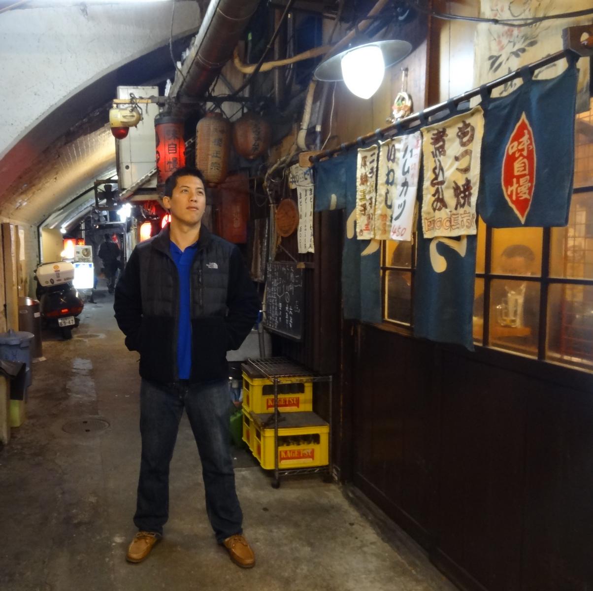 A man looks up in a narrow, atmospheric Tokyo alley lined with lanterns and shop signs.