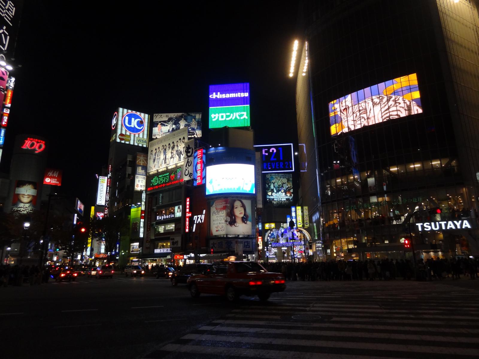 A bustling Tokyo street at night, illuminated by numerous bright neon signs and electronic billboards.