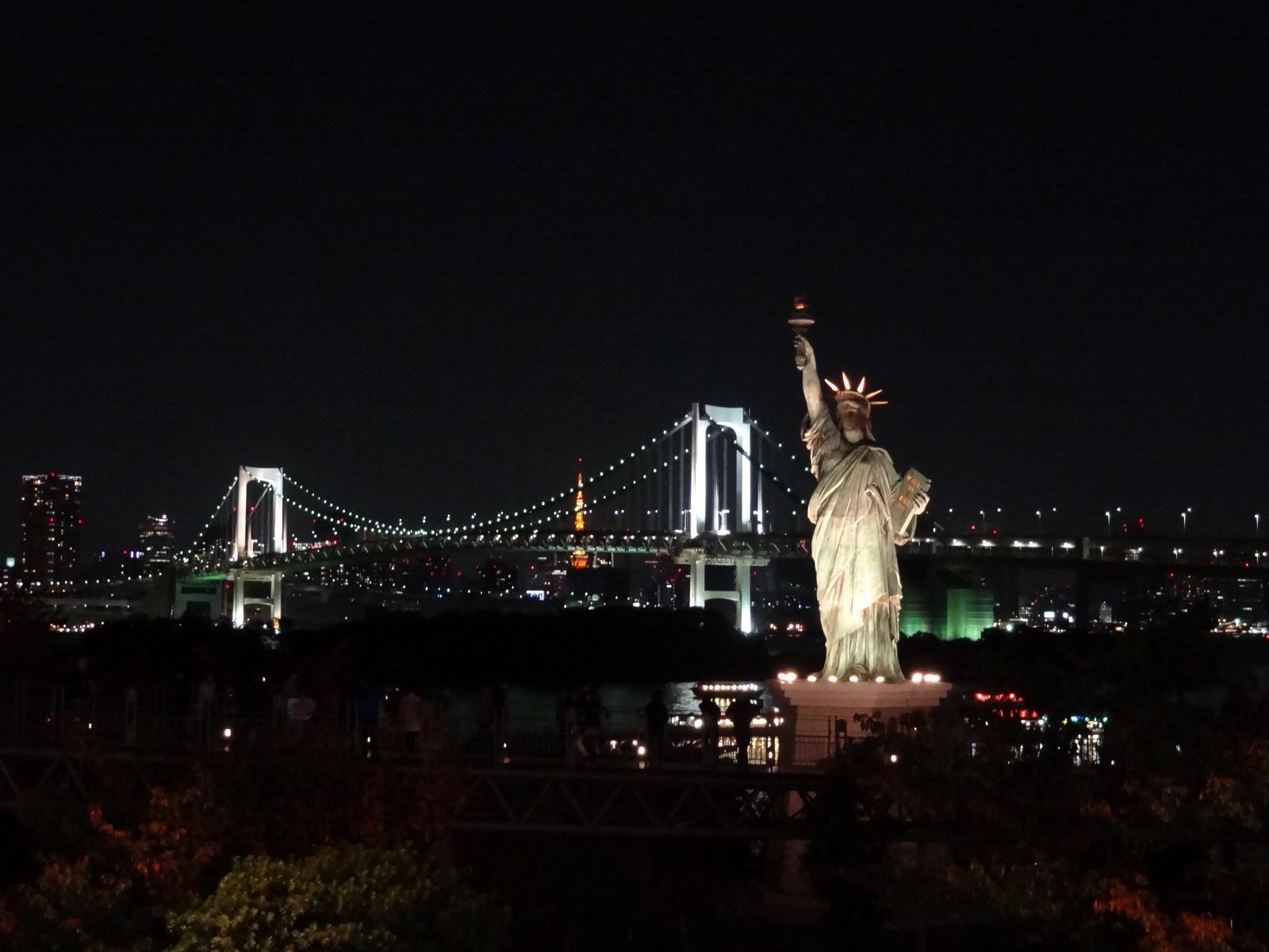 A night view of the Tokyo Statue of Liberty replica in front of the illuminated Rainbow Bridge and Tokyo Tower.