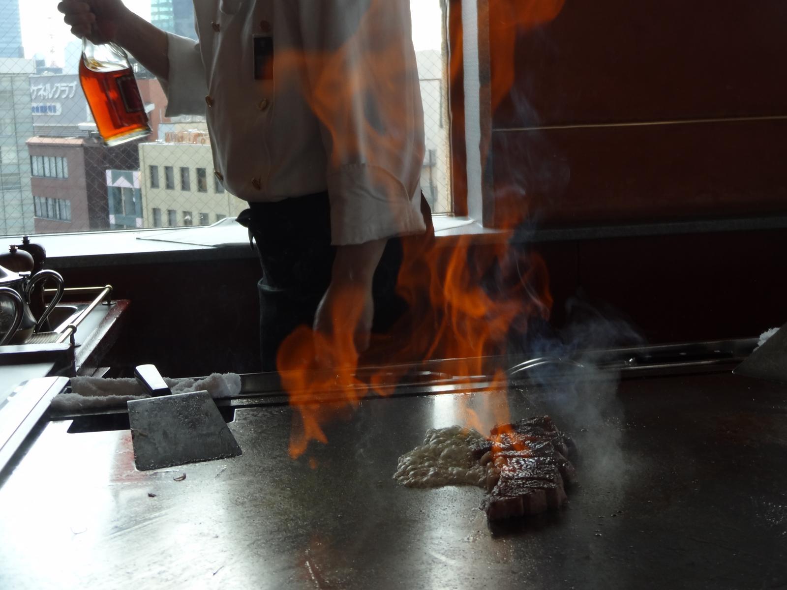 A chef flambés a steak and other food on a teppanyaki grill.
