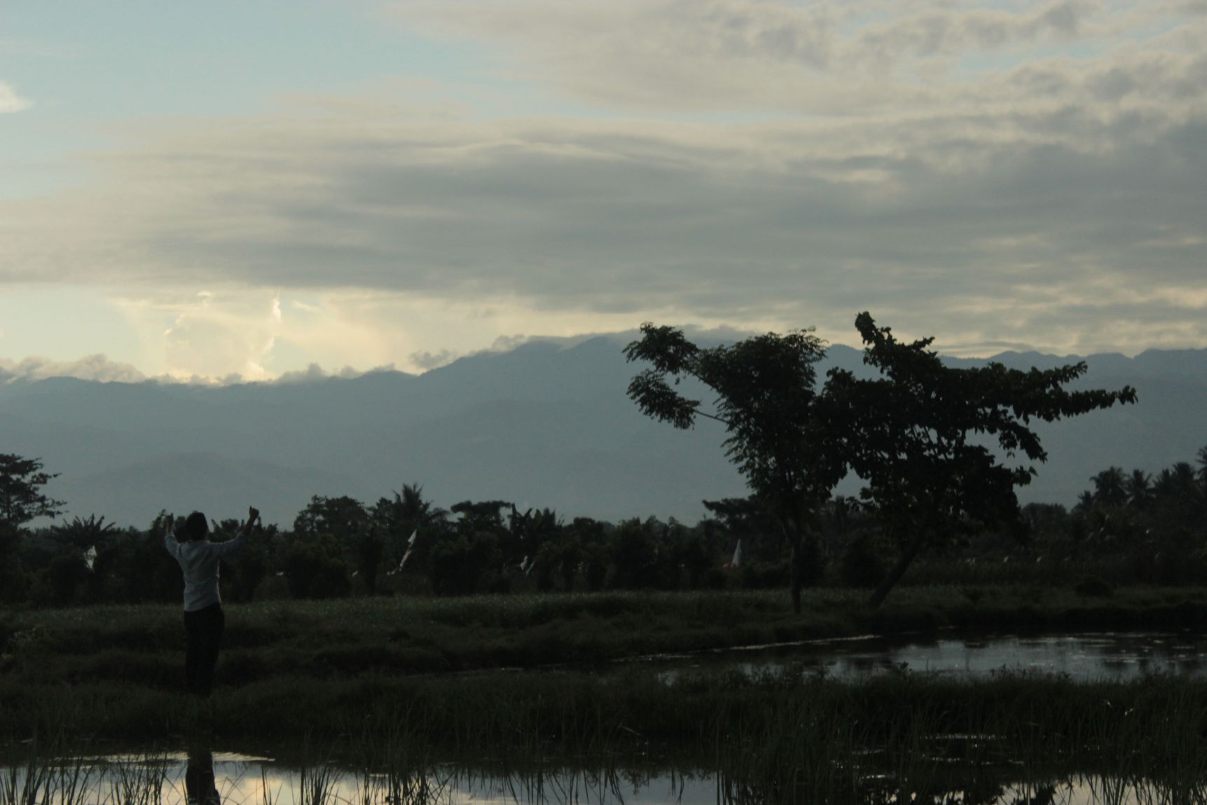 A person stands silhouetted with arms raised by a reflective pool against a cloudy sky and distant mountains, during a total solar eclipse.