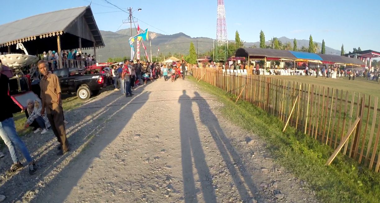 People gather on a dirt path with long shadows, a traditional building, and an event area with tents in Palu, Indonesia.