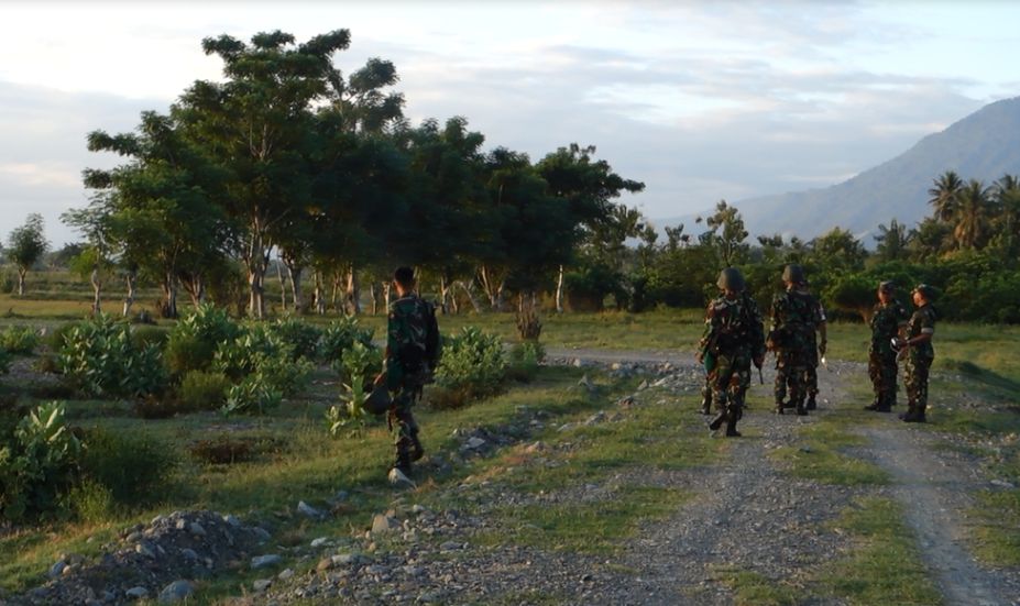 Several soldiers walk on a dirt path through a green, mountainous landscape in Palu, Indonesia.