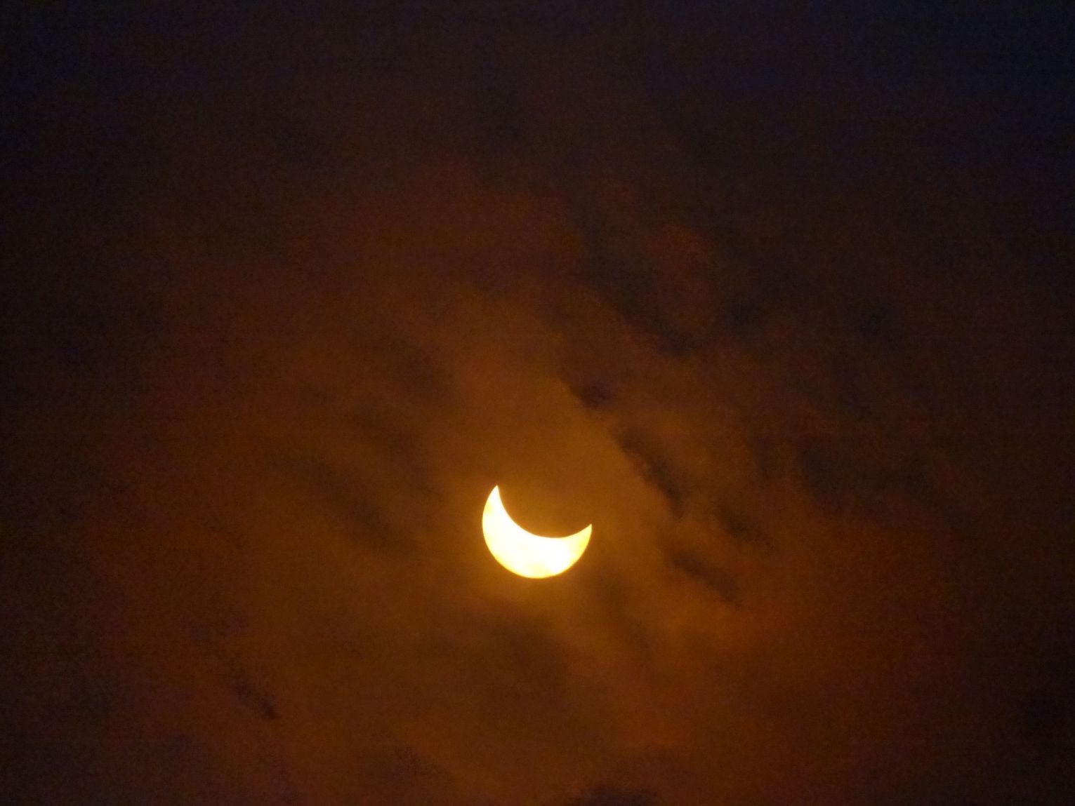 A brilliant crescent sun appears through dark, orange-hued clouds during a solar eclipse.
