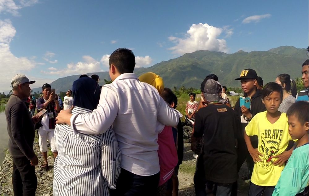 A group of people gather outdoors in Palu, Indonesia, with green mountains and a blue sky in the background.