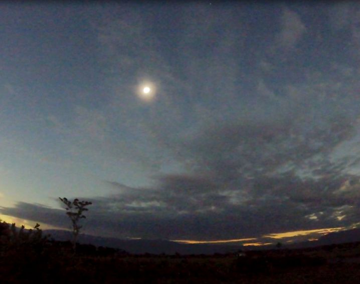 A total solar eclipse with the sun's bright corona visible in a dark, cloudy sky over a shadowed landscape.