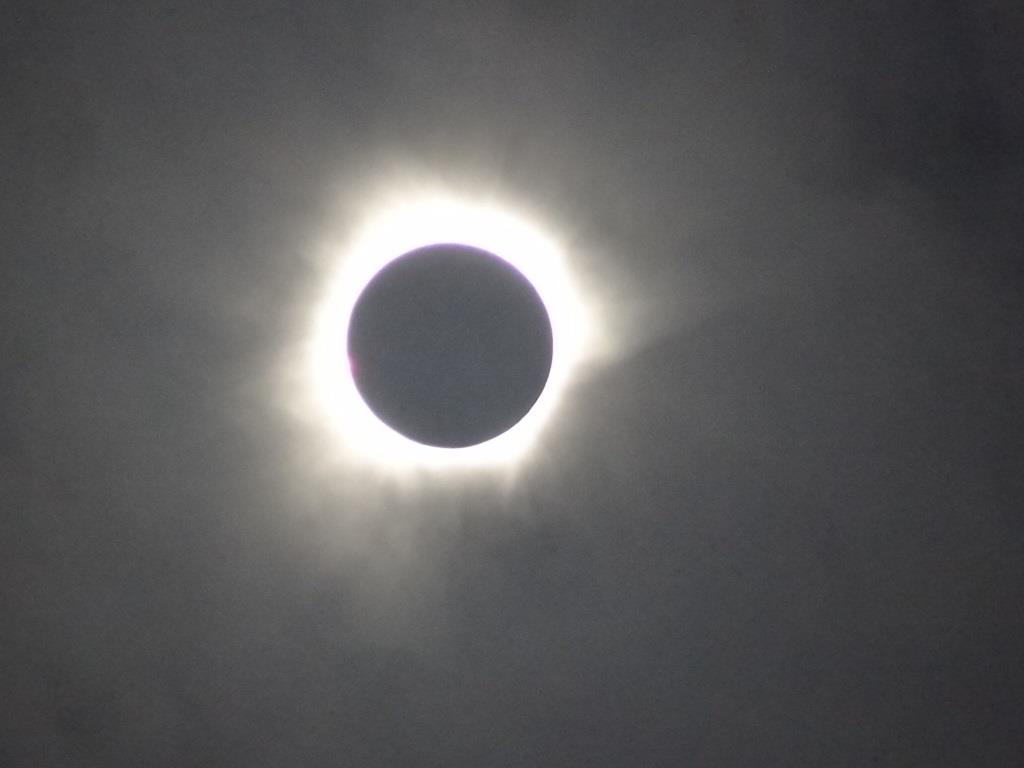 Total solar eclipse with a bright corona visible around the dark disk of the Moon against a cloudy sky.