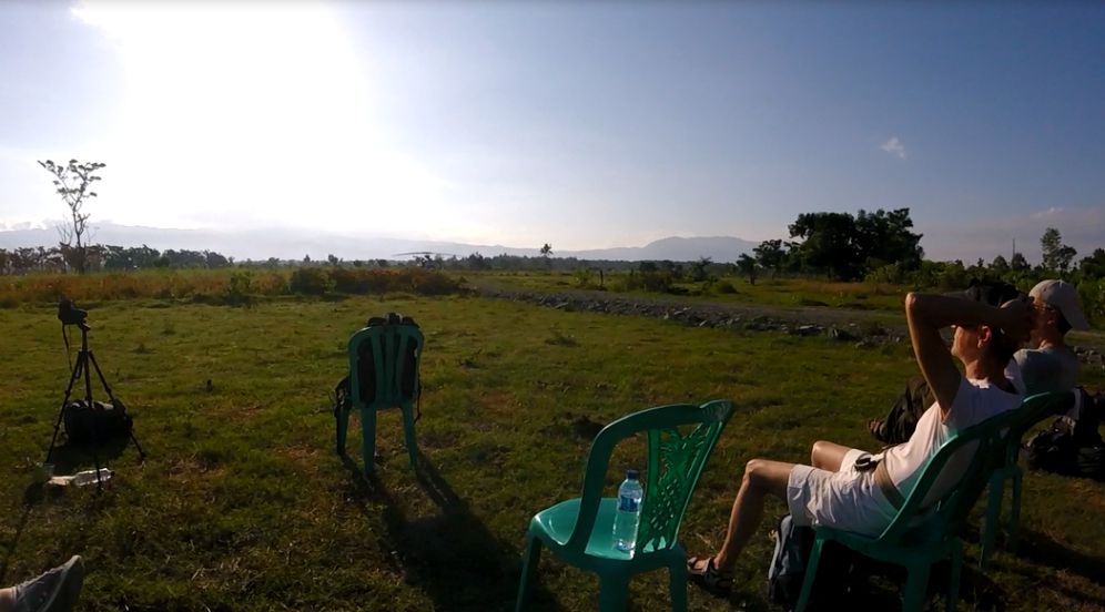 Two people relax in chairs in a grassy field with camera gear set up, looking towards the bright sky, anticipating a total solar eclipse.