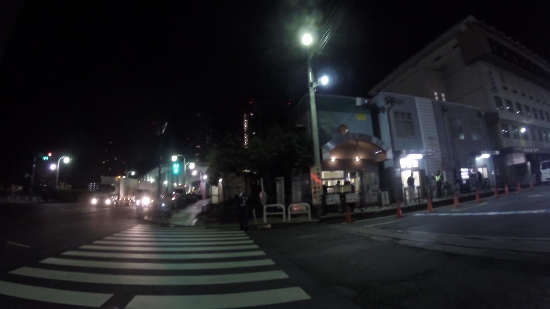 A dark early morning street scene in Tsukiji with a wide pedestrian crossing and buildings illuminated by streetlights.